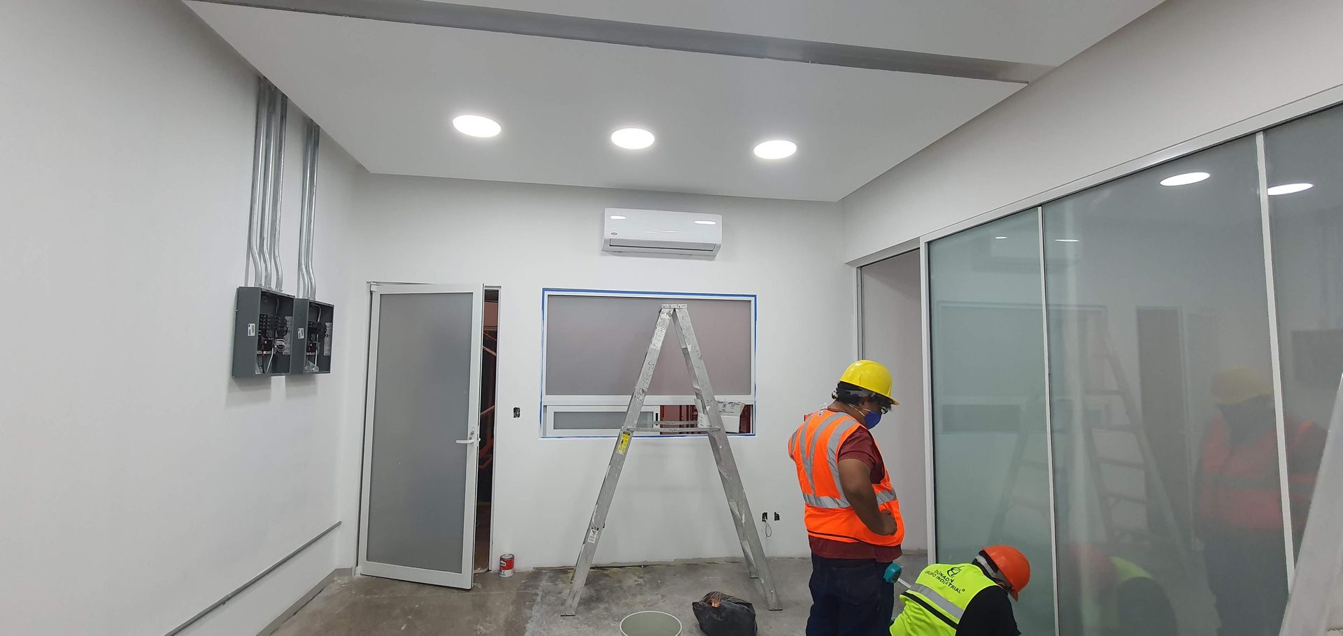 Dos trabajadores con chalecos de seguridad y cascos trabajan dentro de una habitación con una escalera de mano, una mampara de cristal y paneles eléctricos.