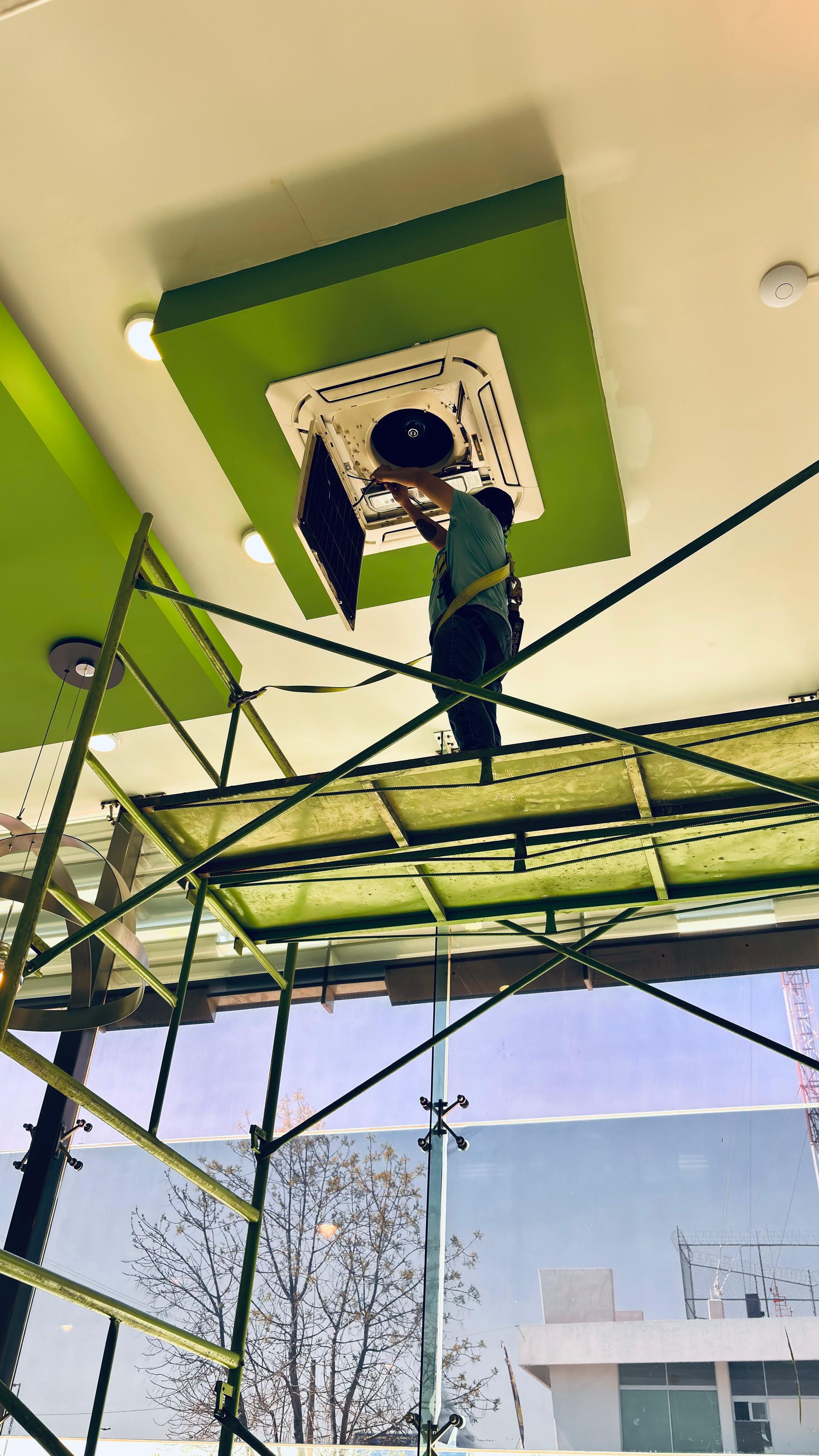 Un trabajador subido a un andamio repara una unidad de aire acondicionado cuadrada de color blanco, montada en el techo y enmarcada en un marco de color verde brillante.