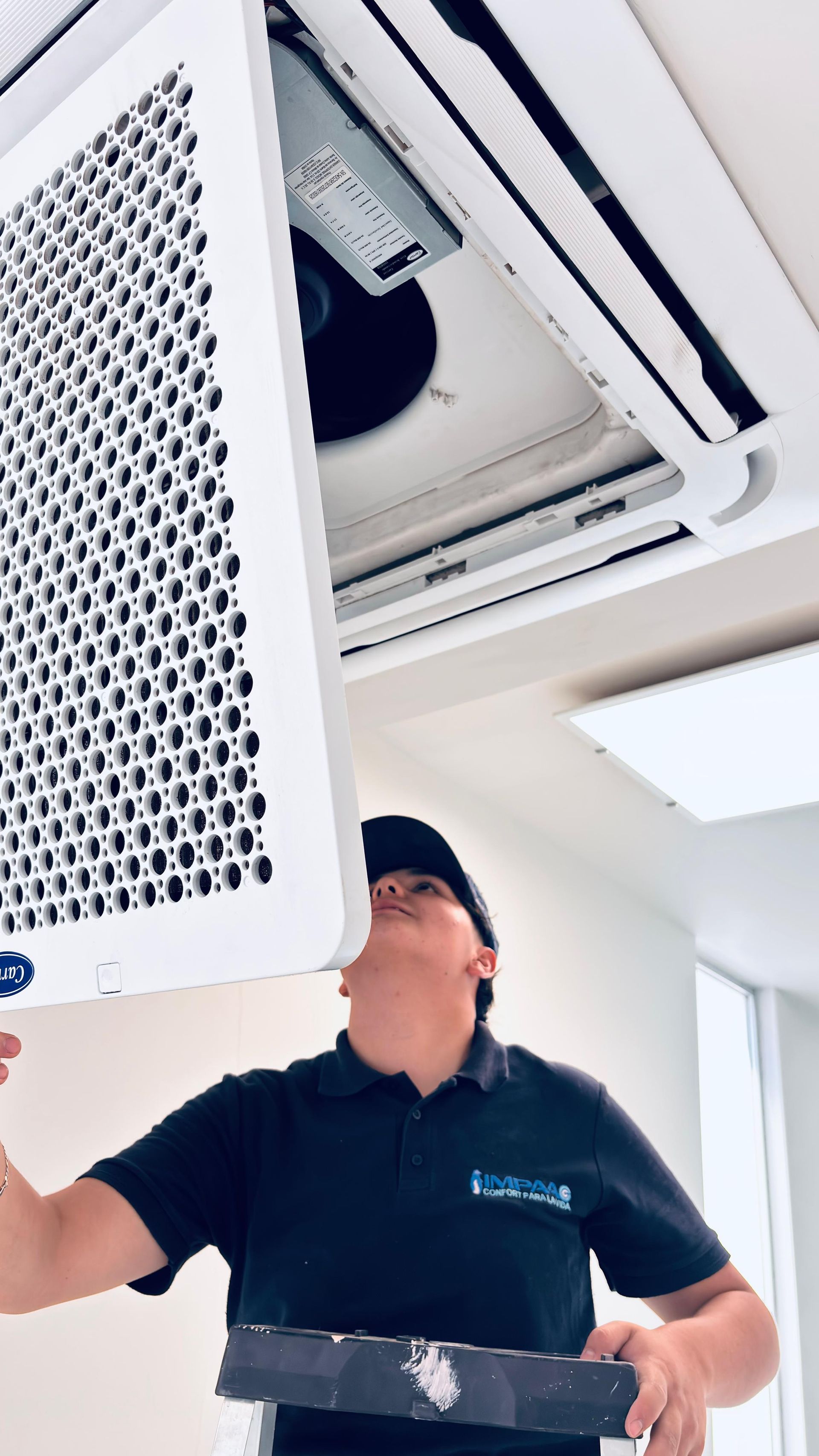 Un técnico de servicio con uniforme negro realiza tareas de mantenimiento en una unidad de aire acondicionado montada en el techo.