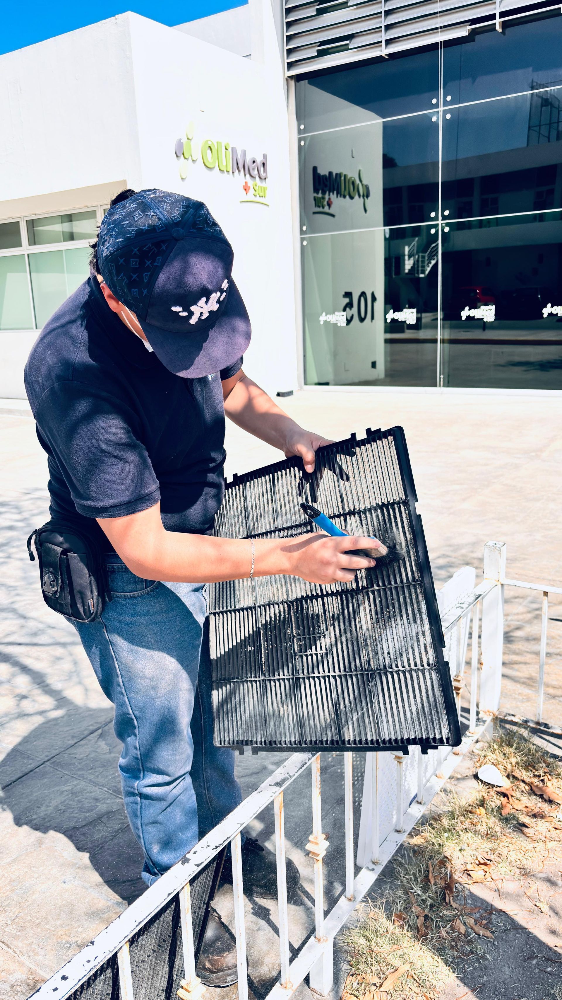 Un técnico con gorro y ropa oscura limpia un filtro de malla negra de un aire acondicionado en el exterior, cerca de la entrada de un edificio.