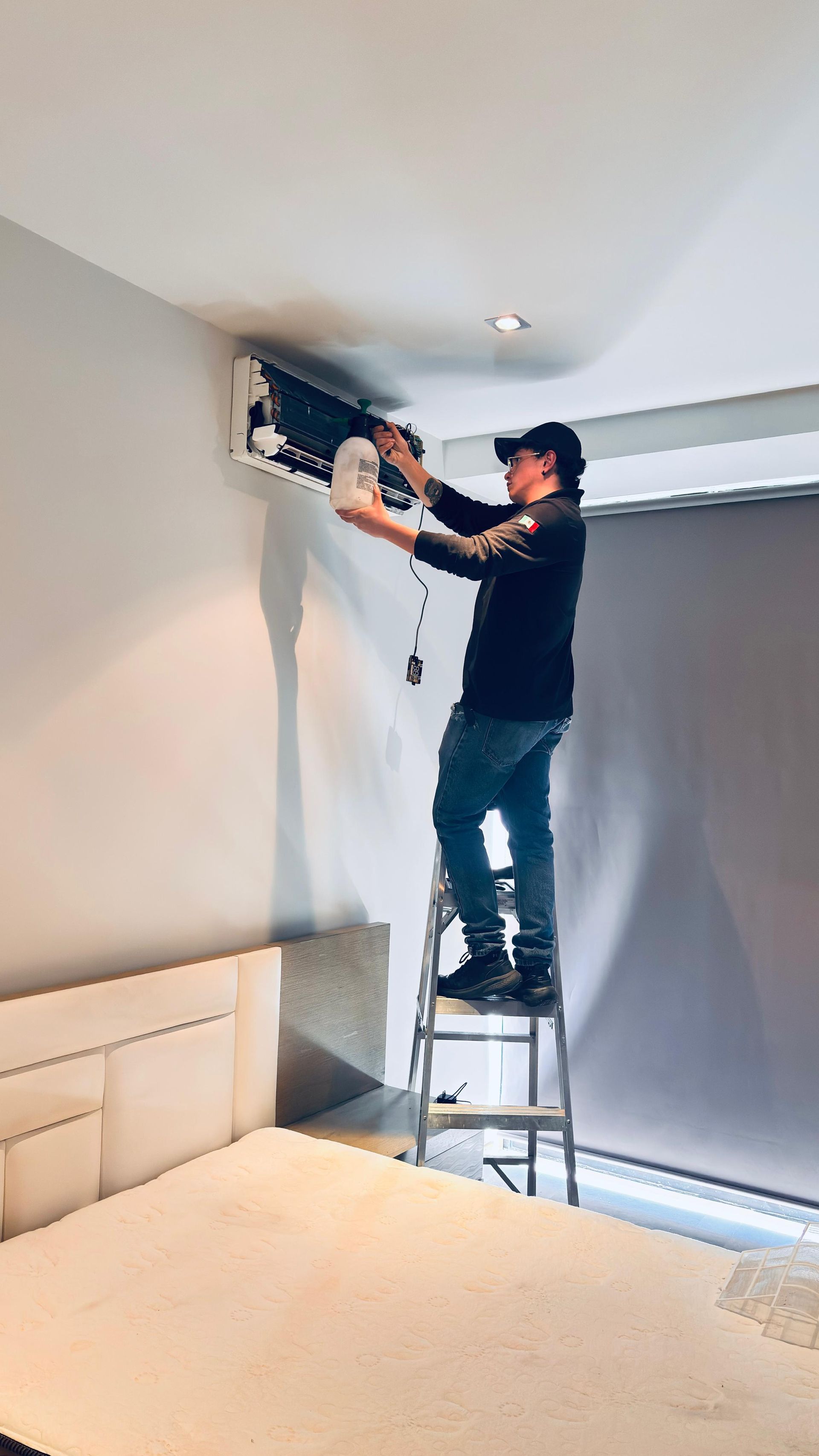 Una persona está subida a una escalera limpiando o reparando una unidad de aire acondicionado montada en la pared de un dormitorio.