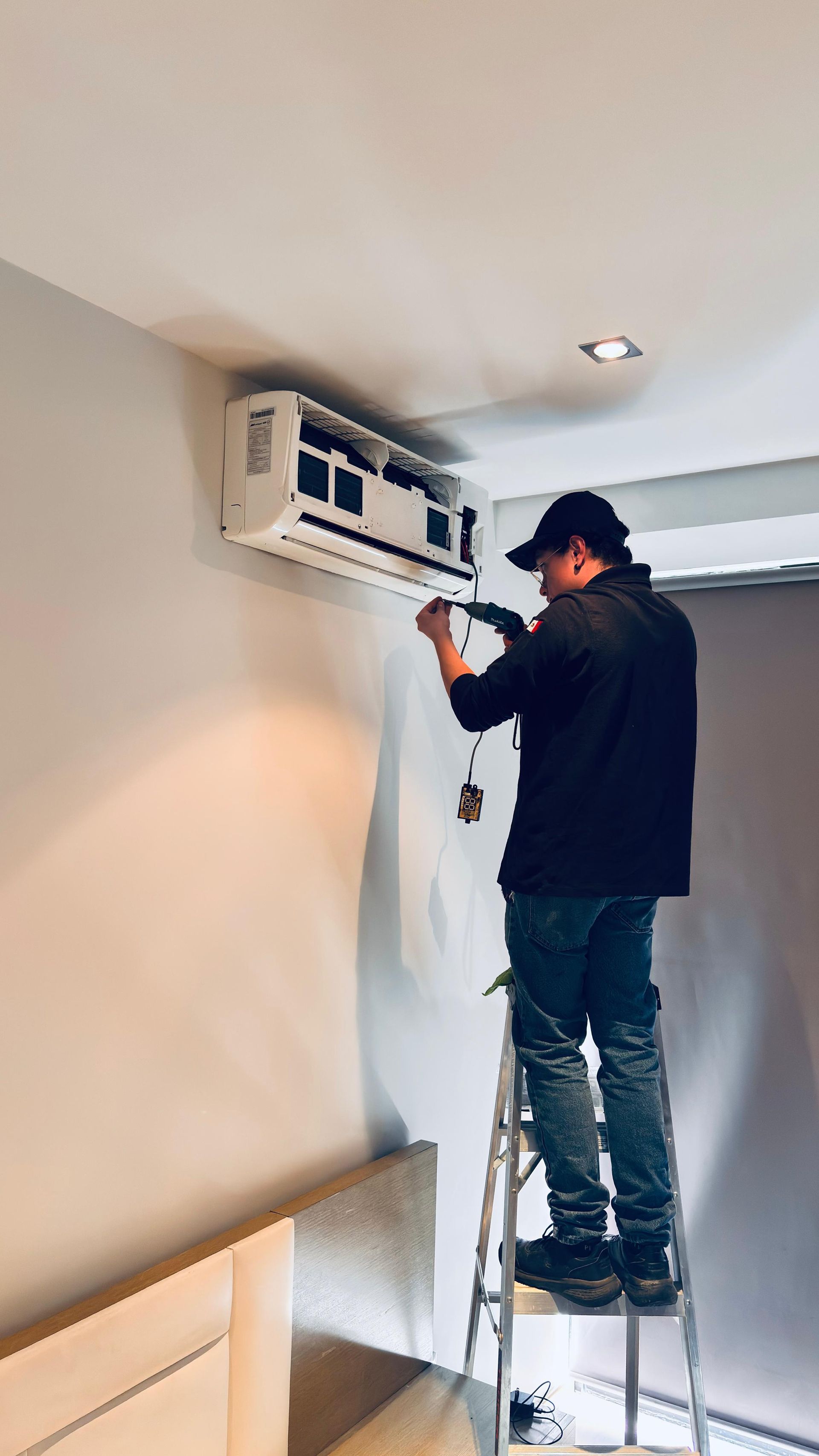 Un técnico se encuentra subido a una escalera, limpiando o reparando un aire acondicionado montado en la pared en una habitación interior bien iluminada.