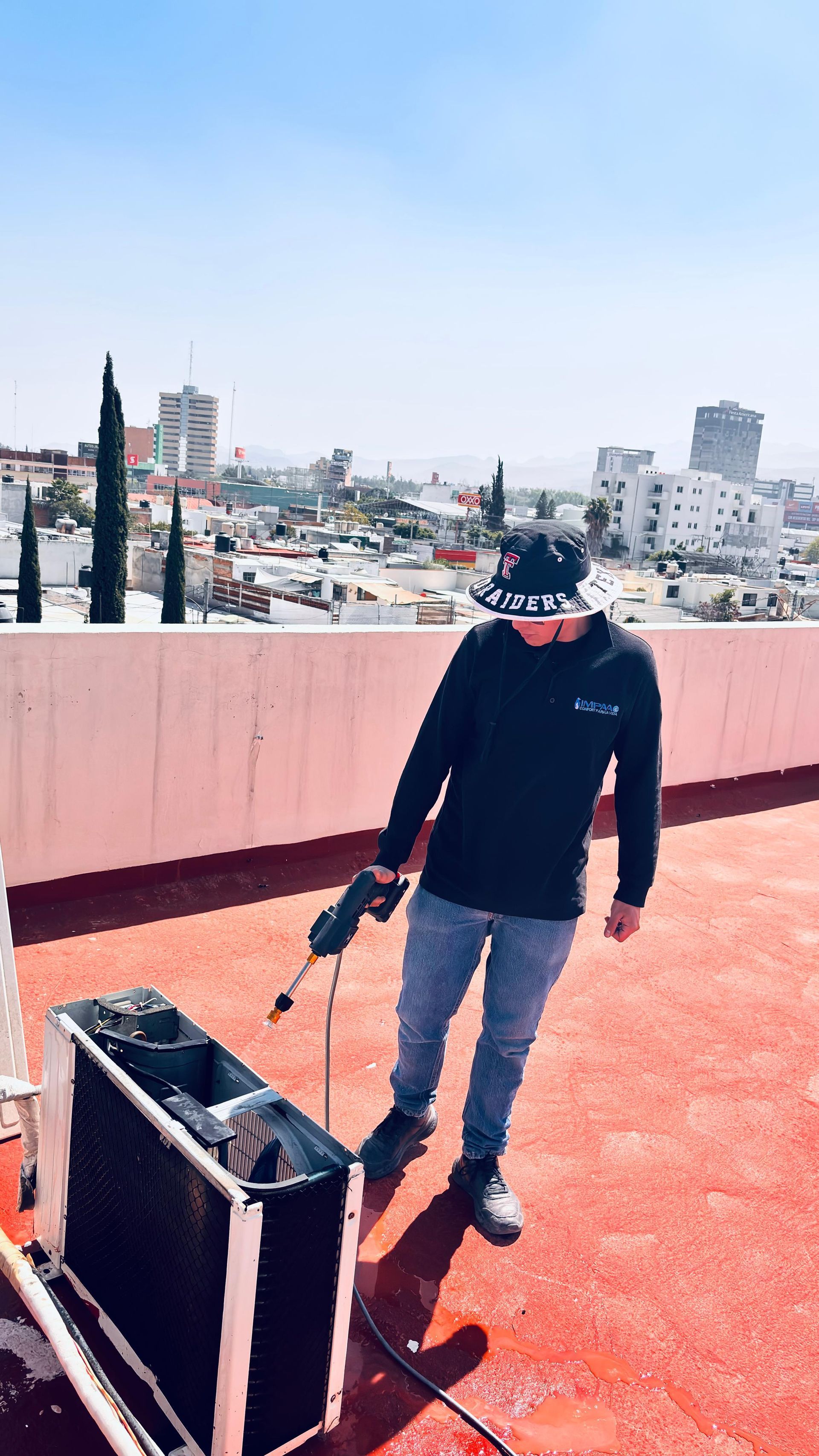 Una persona con sombrero de pescador y ropa oscura utiliza una hidrolimpiadora en una unidad de aire acondicionado exterior en una terraza urbana en la azotea.