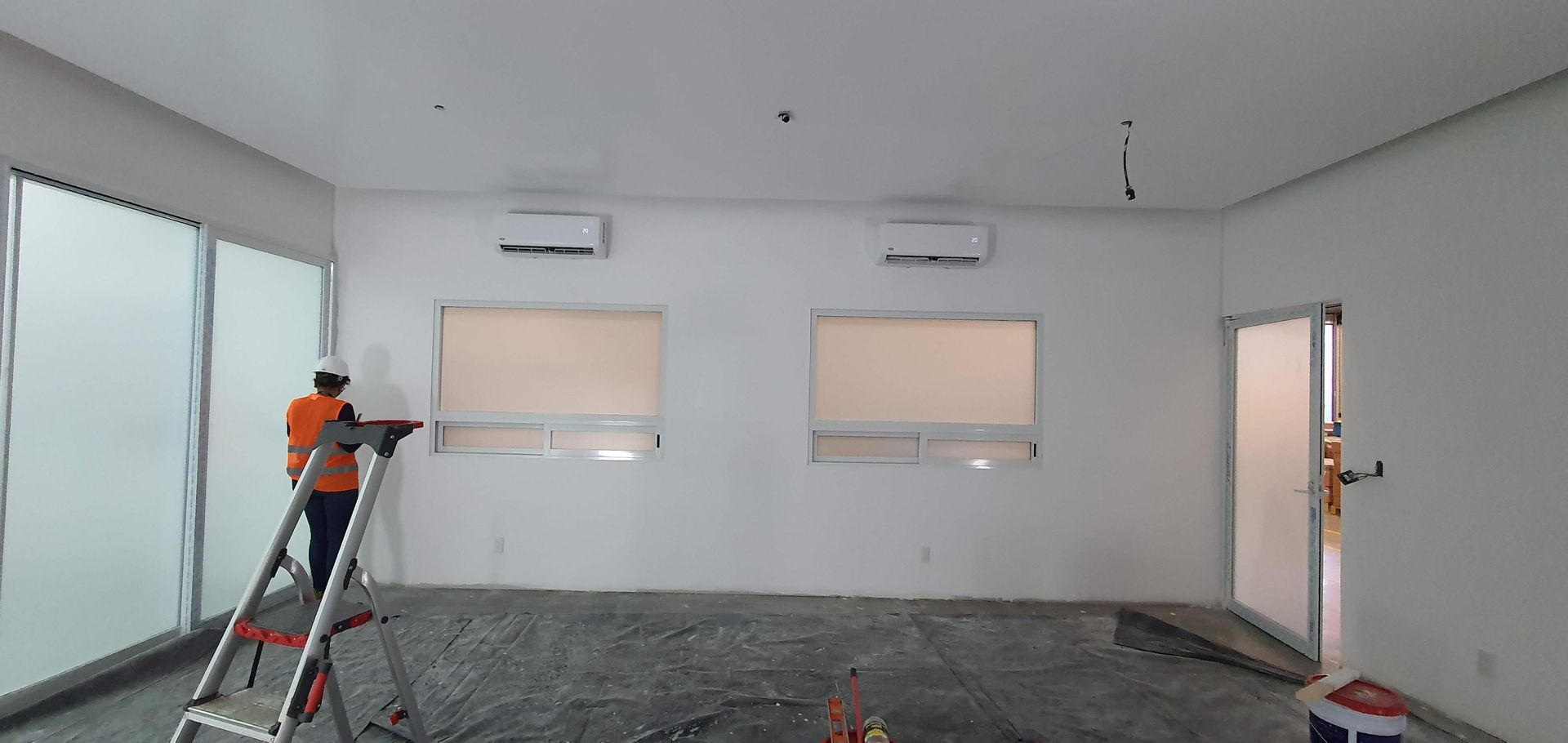 Un trabajador con un chaleco naranja está subido a una escalera en una habitación blanca con dos ventanas, dos unidades de aire acondicionado y una puerta de cristal.