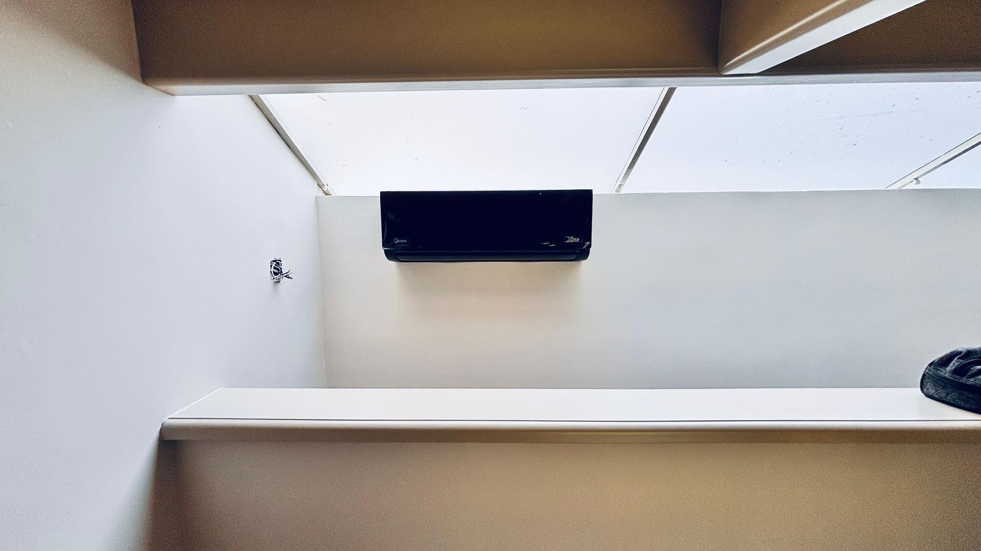 Un aparato de aire acondicionado negro montado sobre una pared blanca en una habitación minimalista con techo inclinado y translúcido.