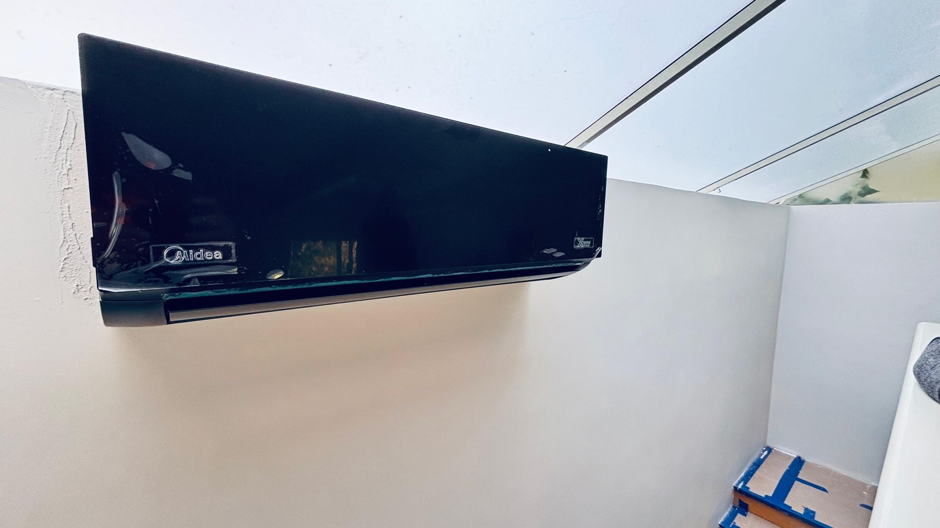 Un aparato de aire acondicionado negro montado en la pared e instalado sobre una pared blanca en un espacio interior.