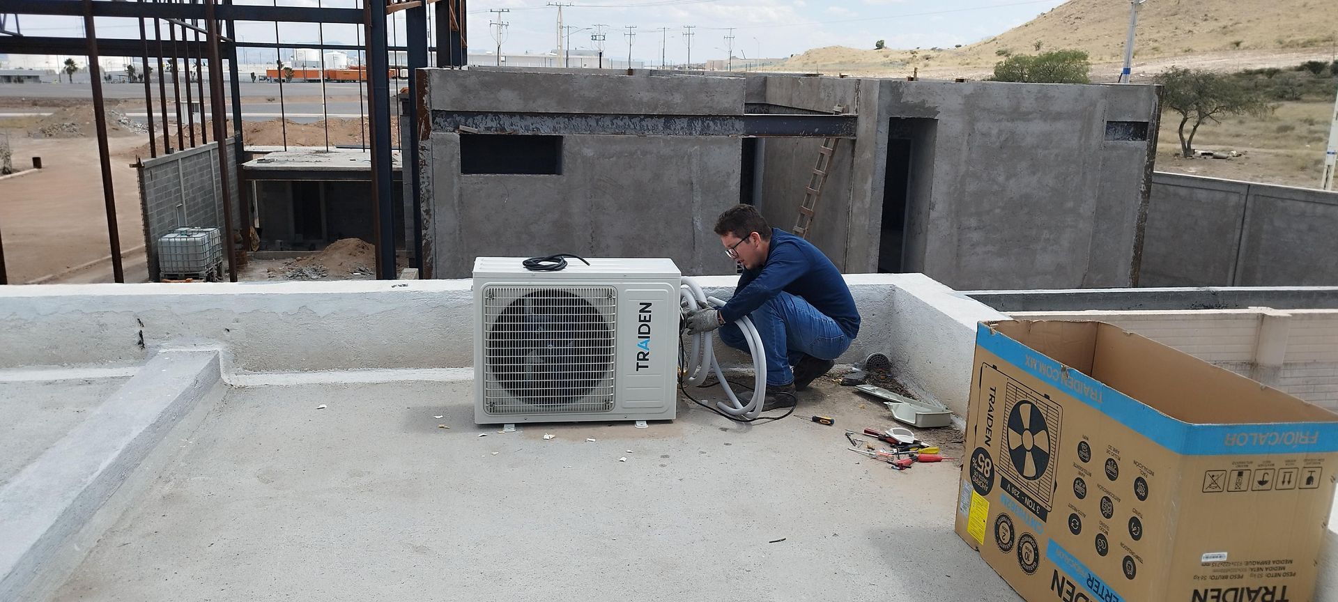 Un técnico instala una unidad de aire acondicionado en una azotea plana de hormigón cerca de un edificio en construcción.