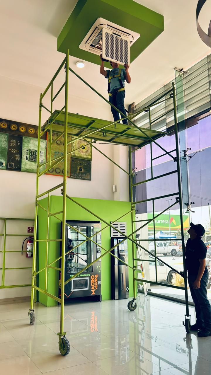 Un técnico se encuentra subido a un andamio alto y verde para realizar el mantenimiento de una unidad de aire acondicionado instalada en el techo de una habitación luminosa.