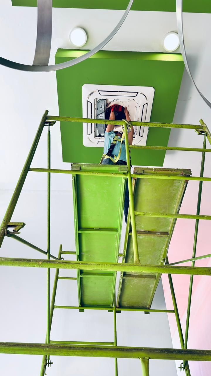Una persona se encuentra sobre un andamio verde, trabajando en una unidad de aire acondicionado blanca montada en el techo de una habitación con detalles en verde.