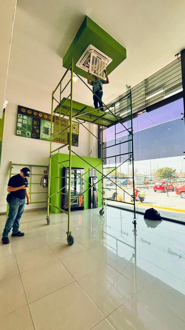 Una persona se encuentra sobre un andamio móvil para instalar o realizar el mantenimiento de una unidad de aire acondicionado montada en el techo dentro de una tienda.