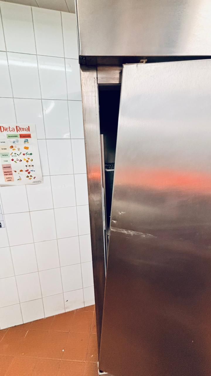 La puerta de un frigorífico comercial de acero inoxidable está ligeramente entreabierta en una cocina con paredes y suelo de baldosas blancas.