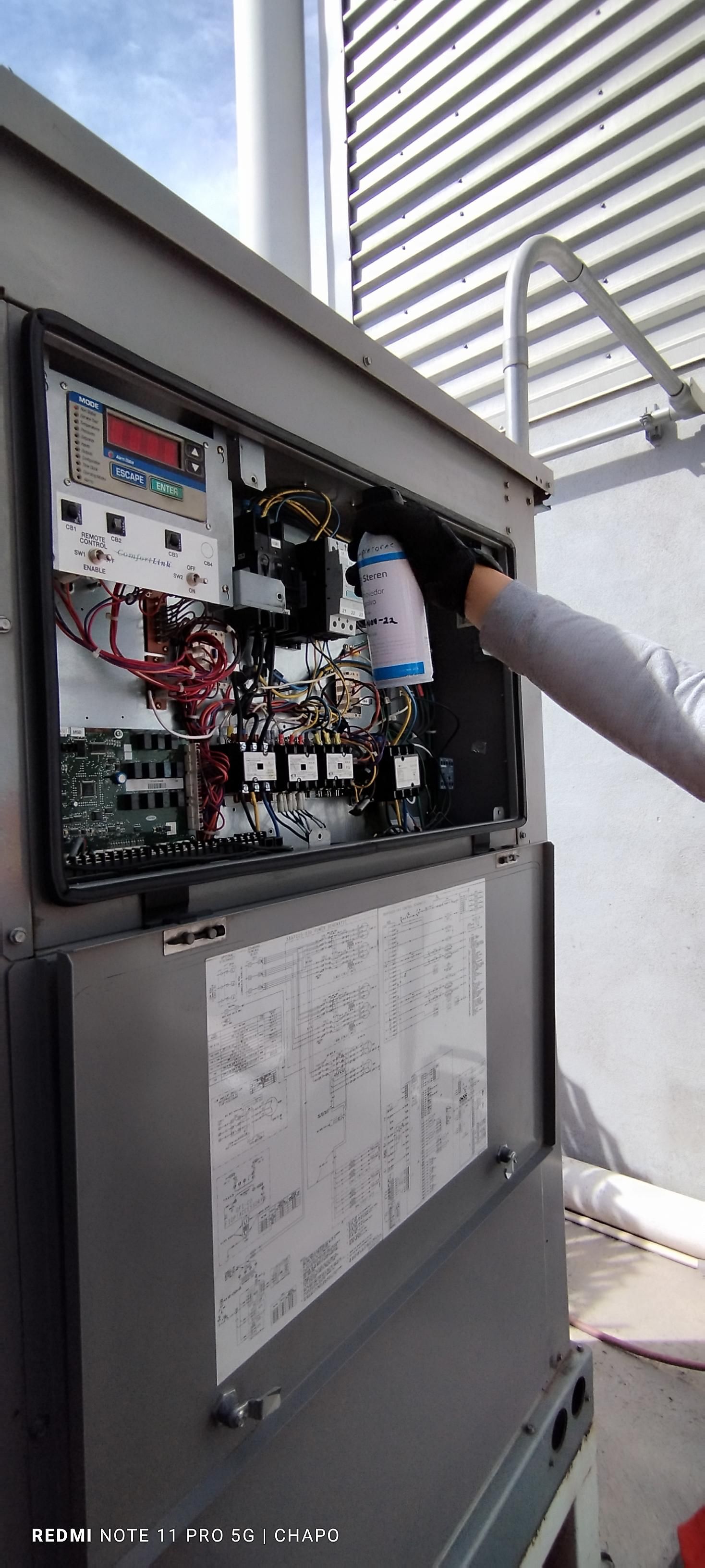 Una persona con mangas grises introduce la mano en un panel eléctrico abierto de una unidad de climatización en la azotea para realizar tareas de mantenimiento.