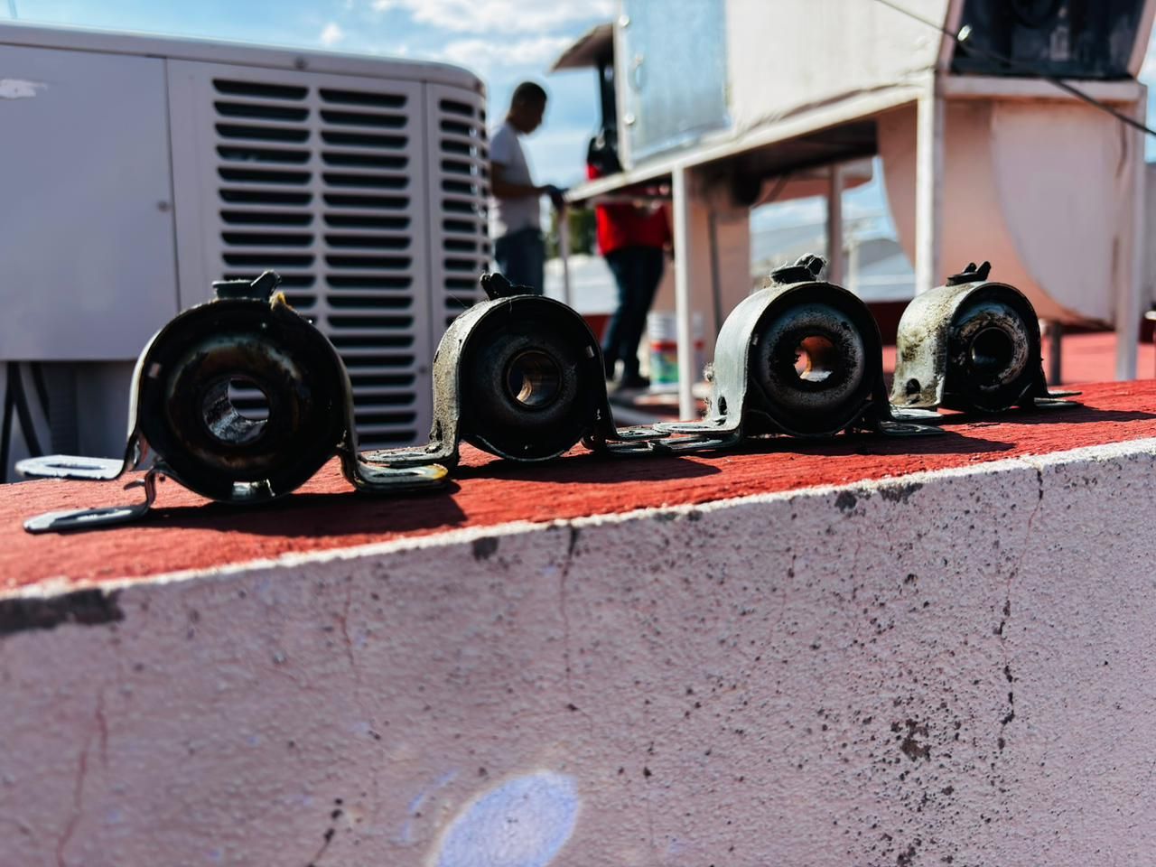 Cuatro casquillos de suspensión usados, de metal y goma, estaban alineados sobre una repisa roja, con gente trabajando al fondo.