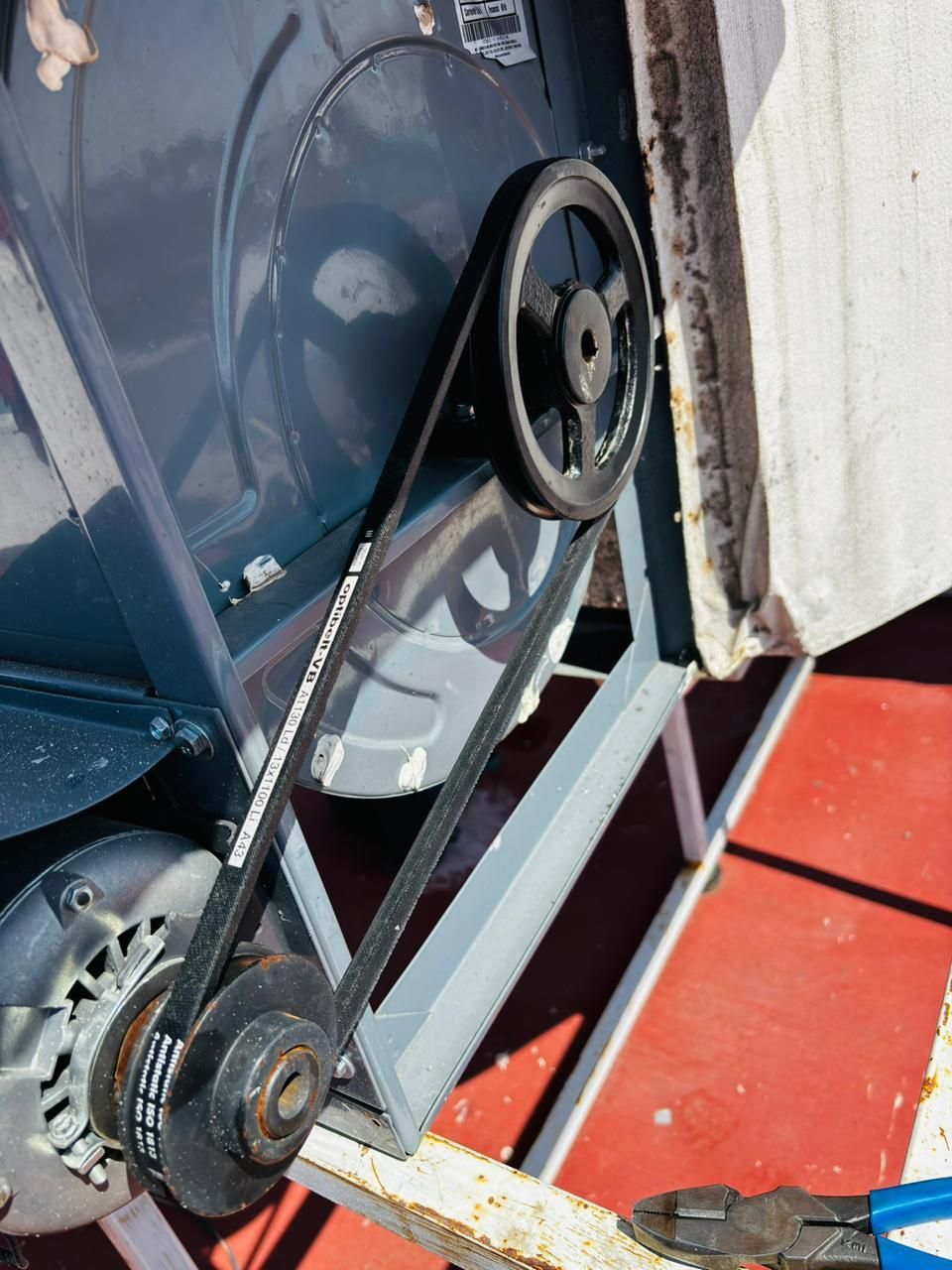 Una correa de transmisión conecta la polea del motor con la polea del ventilador dentro de una unidad de climatización, y en primer plano se aprecian unos alicates.