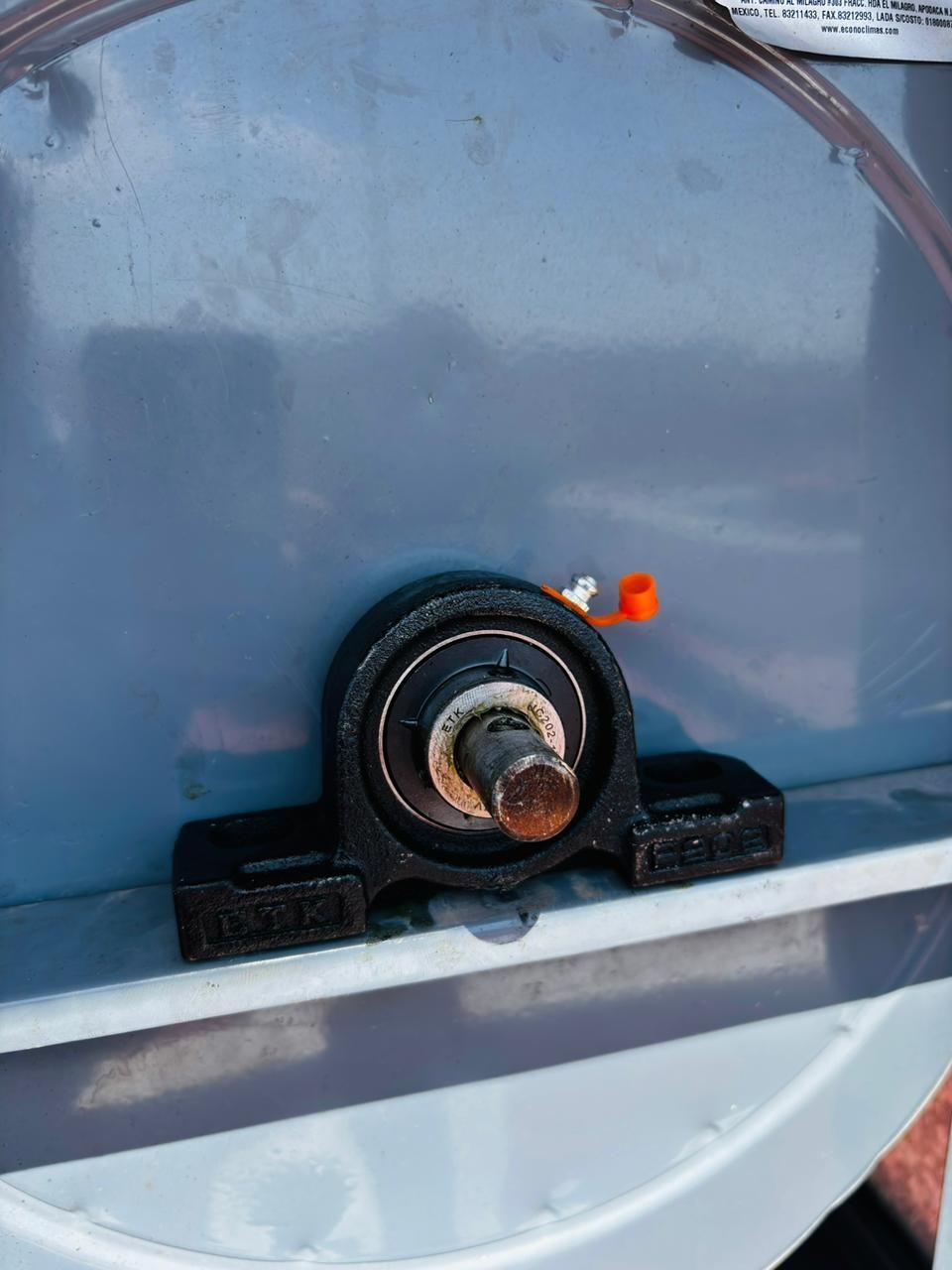 Un cojinete de bloque de metal negro montado sobre una superficie gris con un eje de acero que sobresale y una boquilla de engrase naranja.