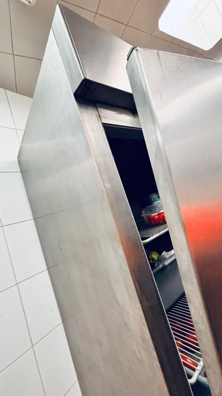 Un frigorífico comercial de acero inoxidable se encuentra apoyado contra una pared de azulejos, con la puerta ligeramente entreabierta.