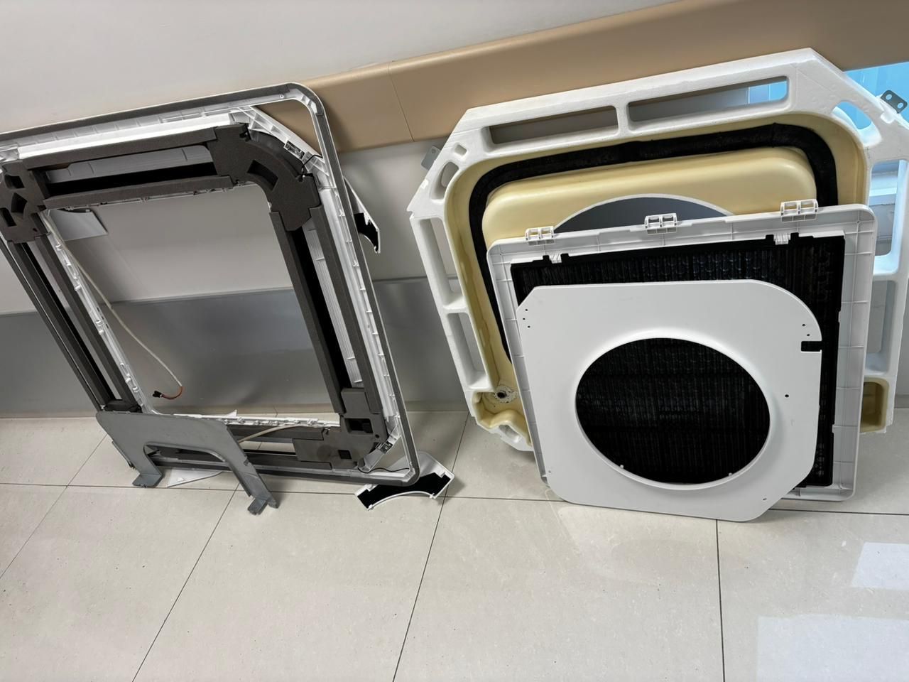 Dos componentes desmontados de una unidad de aire acondicionado de techo, de color blanco y negro, descansan sobre un suelo de baldosas.