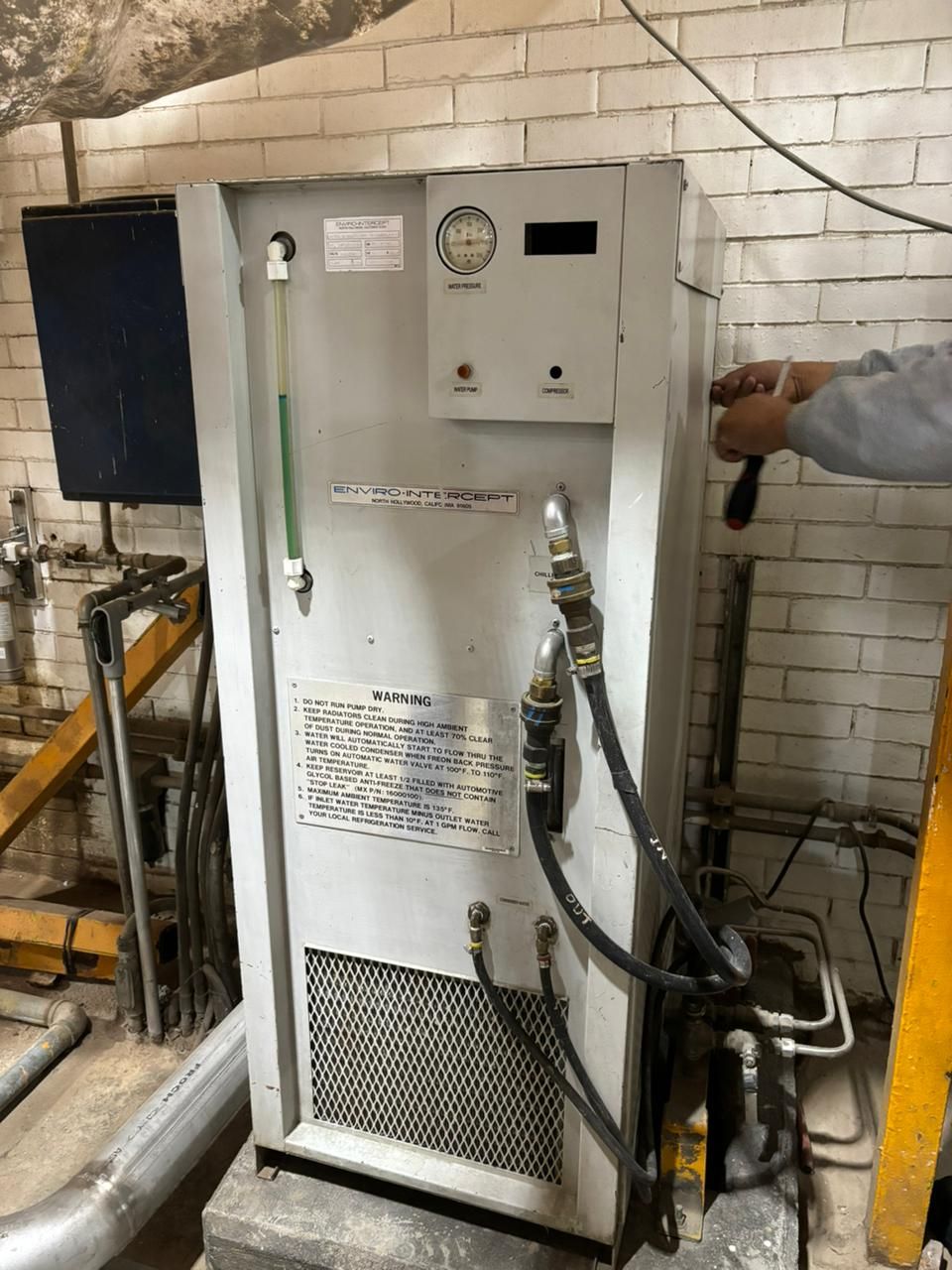 Un trabajador utiliza una herramienta en una secadora industrial alta y gris, que se encuentra sobre una base de hormigón junto a una pared de ladrillo blanco.