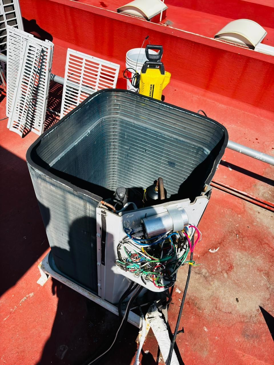 Una unidad condensadora de climatización exterior abierta, con serpentines y cableado expuestos, situada sobre un tejado rojo al que se le han retirado los paneles metálicos.