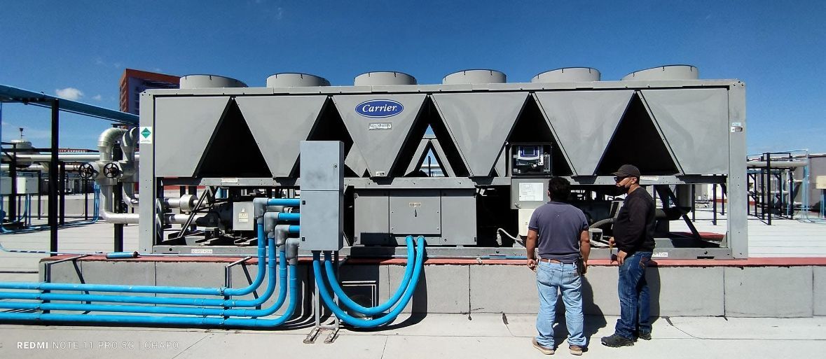 Dos personas están de pie en una azotea junto a una gran unidad de refrigeración industrial por aire, con tuberías azules y un cielo azul de fondo.