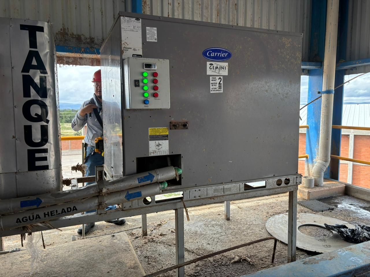 Una unidad de refrigeración industrial Carrier con tuberías etiquetadas se encuentra sobre una plataforma, con un trabajador visible al fondo.