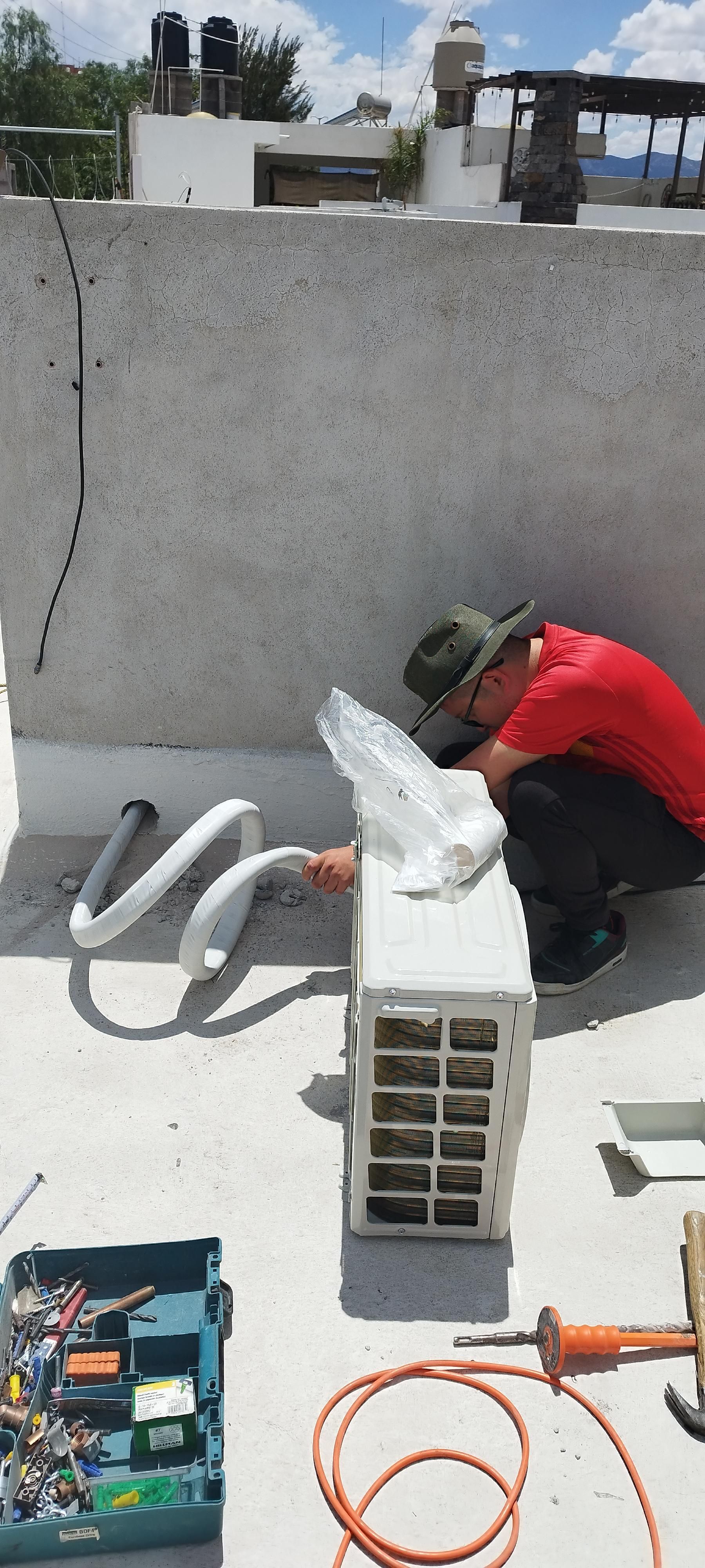 Una persona con camisa y gorra rojas está arrodillada en una azotea blanca, trabajando en una unidad de aire acondicionado con herramientas a mano.
