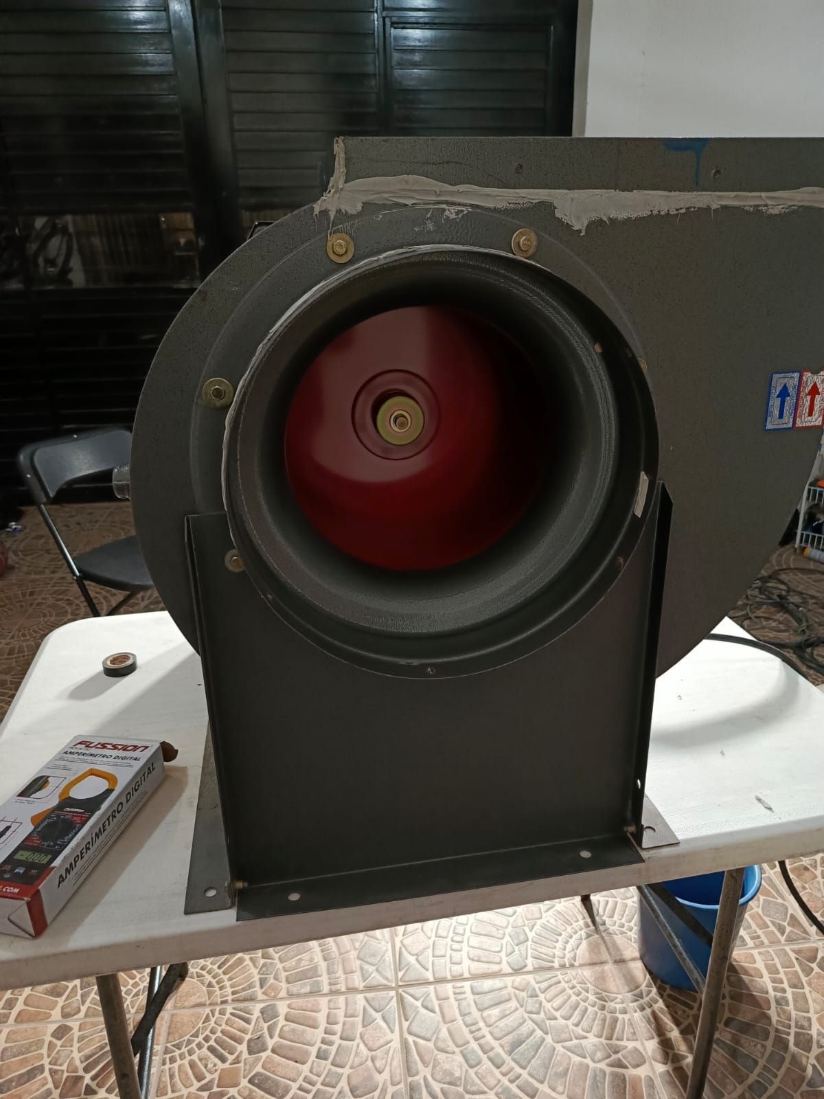 Un ventilador centrífugo industrial gris con una rueda interna roja, colocado sobre una mesa blanca en el interior de una vivienda.