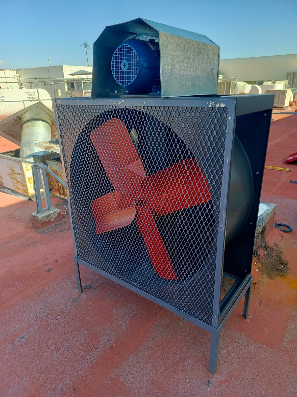 Un ventilador extractor axial industrial con aspas rojas detrás de una rejilla de seguridad, situado en una azotea plana bajo un cielo azul.