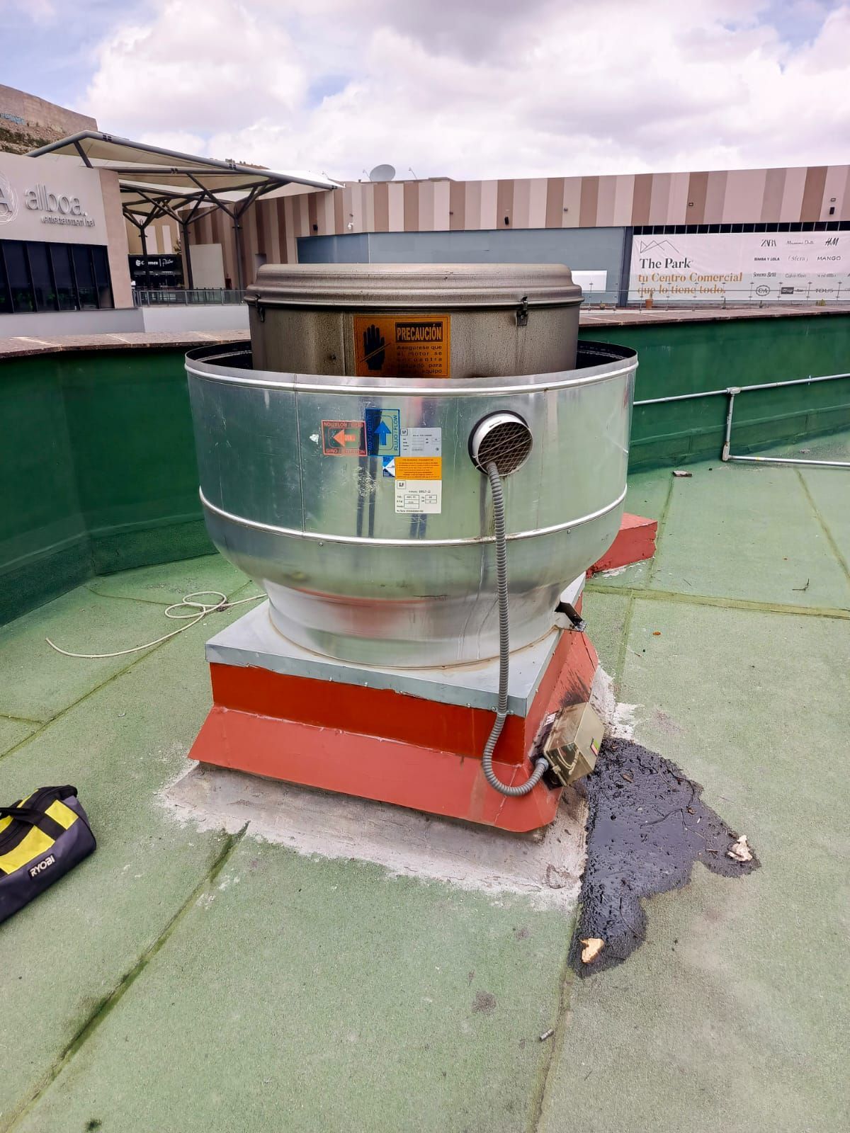 Un extractor de aire metálico instalado en el tejado, sobre un bordillo rojo, con una mancha de sellador gris para techos sobre una superficie verde.