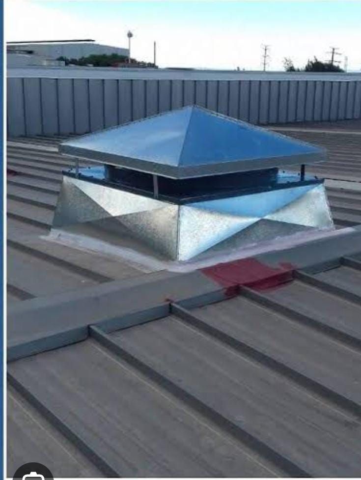 Una campana extractora metálica con forma de pirámide se alza sobre un techo metálico industrial acanalado bajo un cielo brillante.