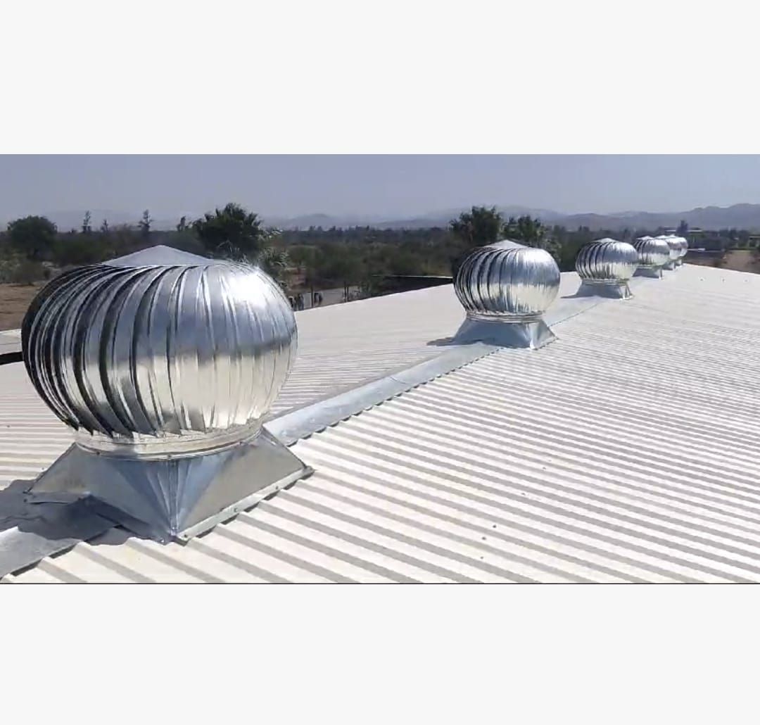 Una hilera de ventiladores de techo metálicos accionados por el viento, instalados en un techo de chapa ondulada blanca bajo un cielo despejado.