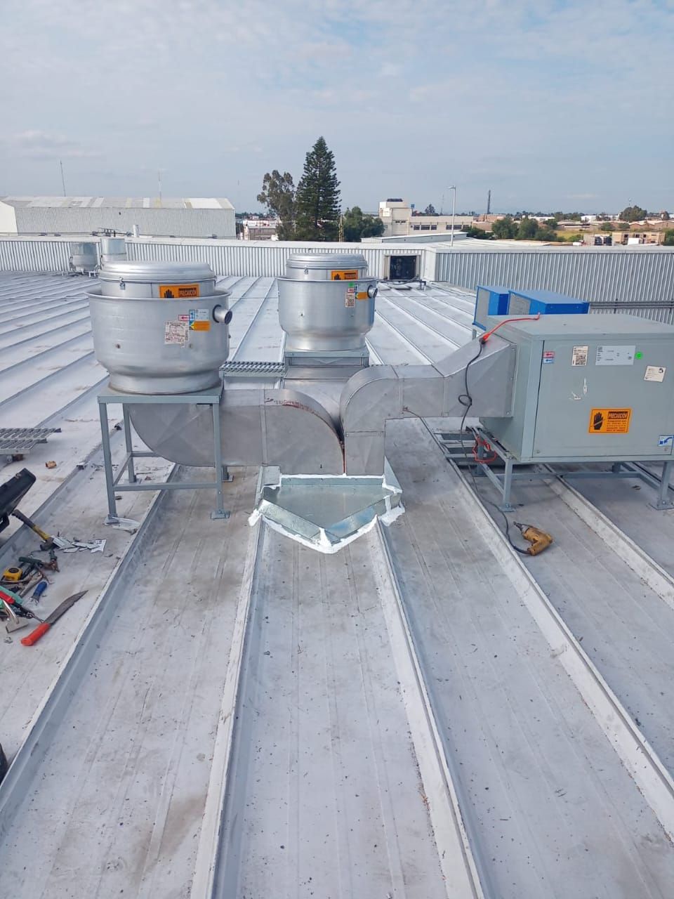 Dos grandes ventiladores industriales de extracción, conectados por conductos, se asientan sobre un techo de chapa ondulada bajo un cielo brillante.
