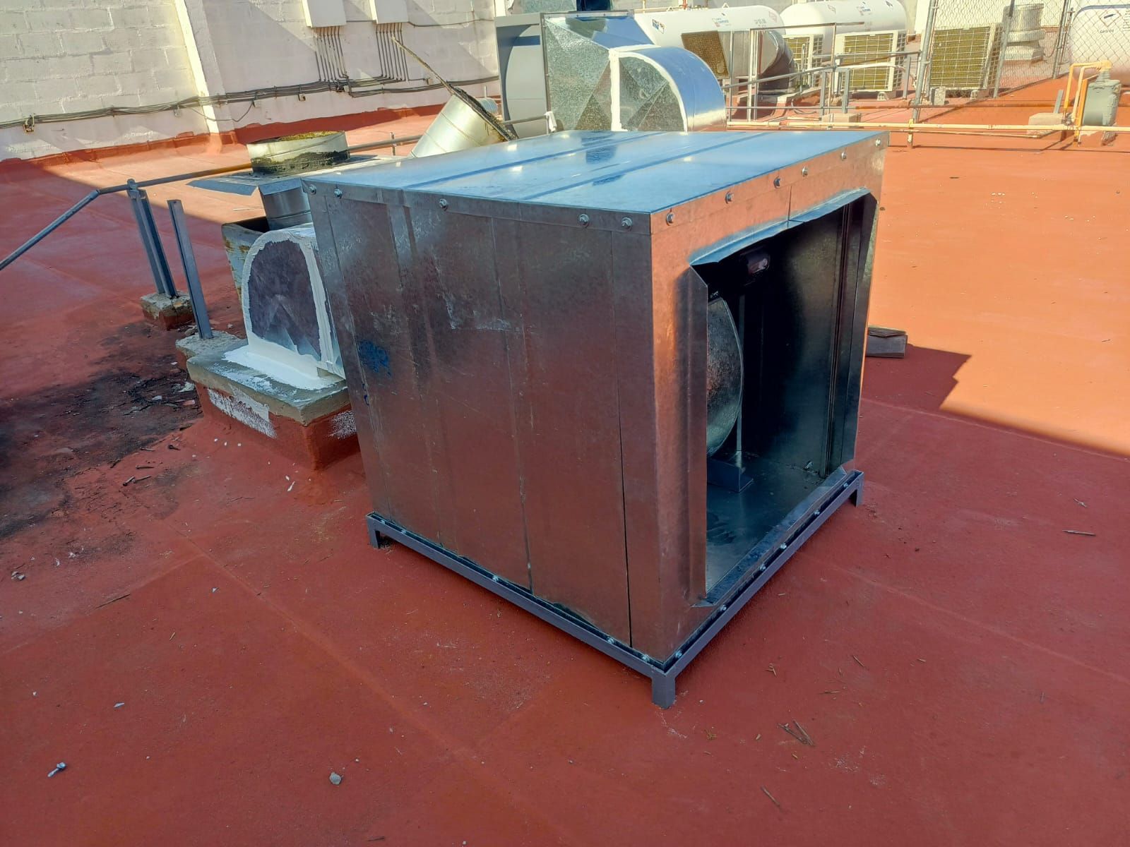 La carcasa metálica de un ventilador centrífugo de climatización se encuentra sobre un tejado plano de color naranja rojizo.