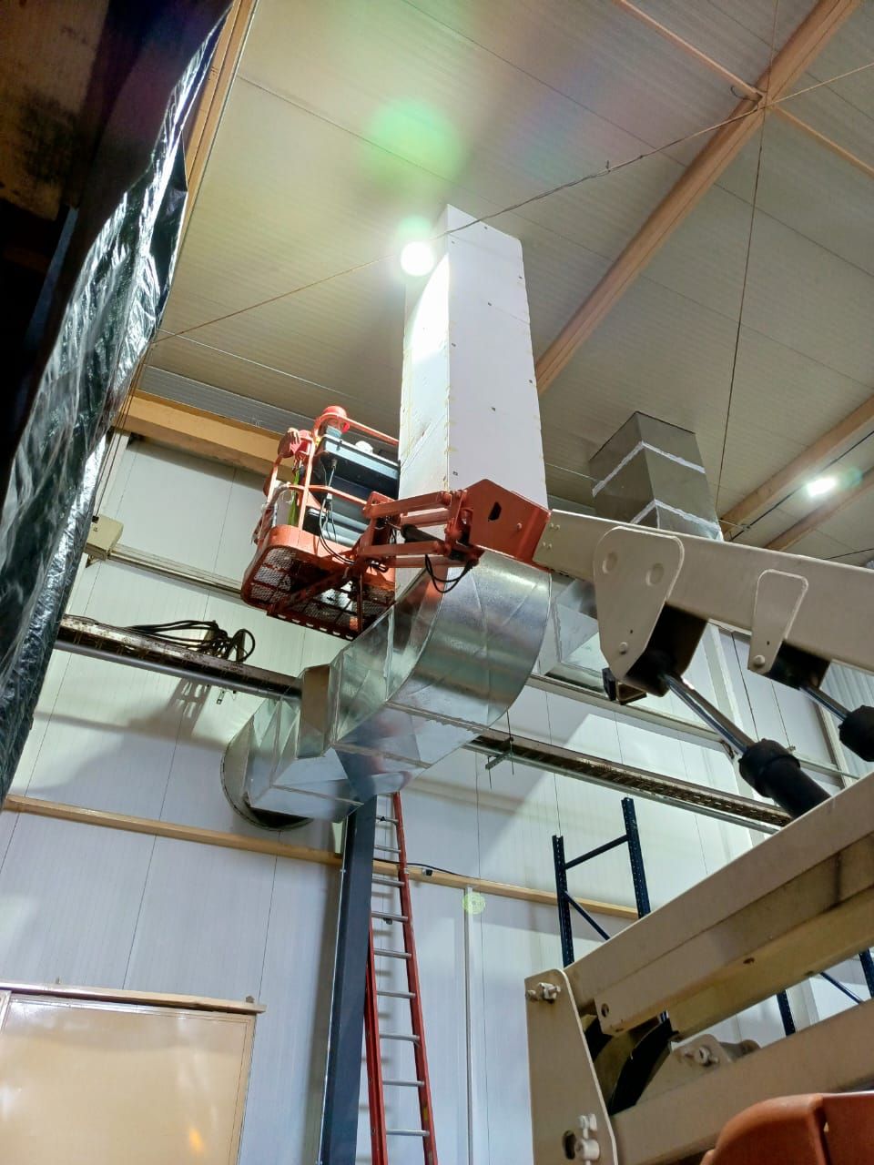 Una plataforma elevadora naranja se encuentra elevada en el interior de un gran edificio, con un trabajador en la cesta instalando conductos metálicos.
