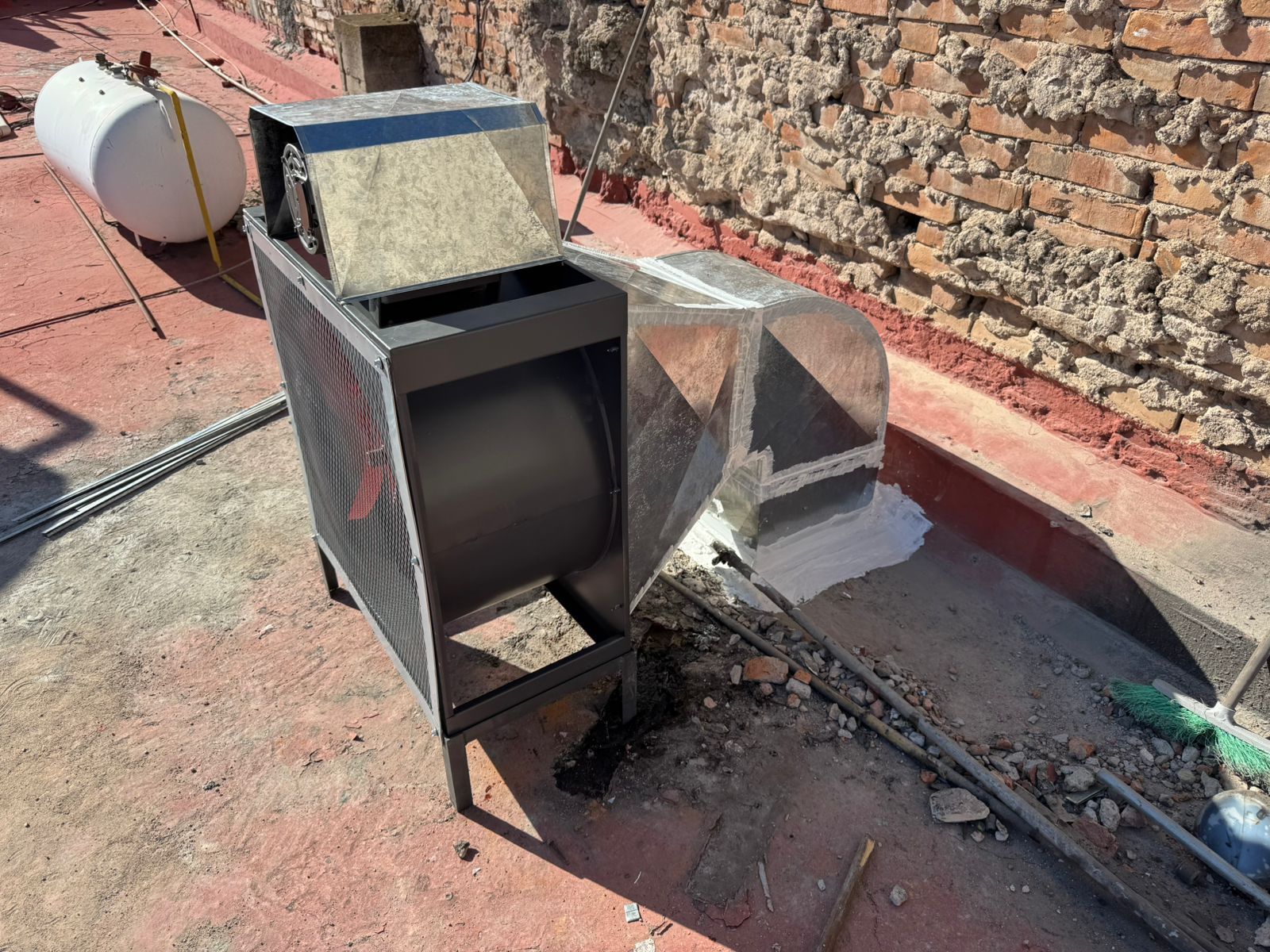 Un ventilador centrífugo industrial de metal se encuentra en una azotea junto a una pared de ladrillos y un tanque cilíndrico blanco.