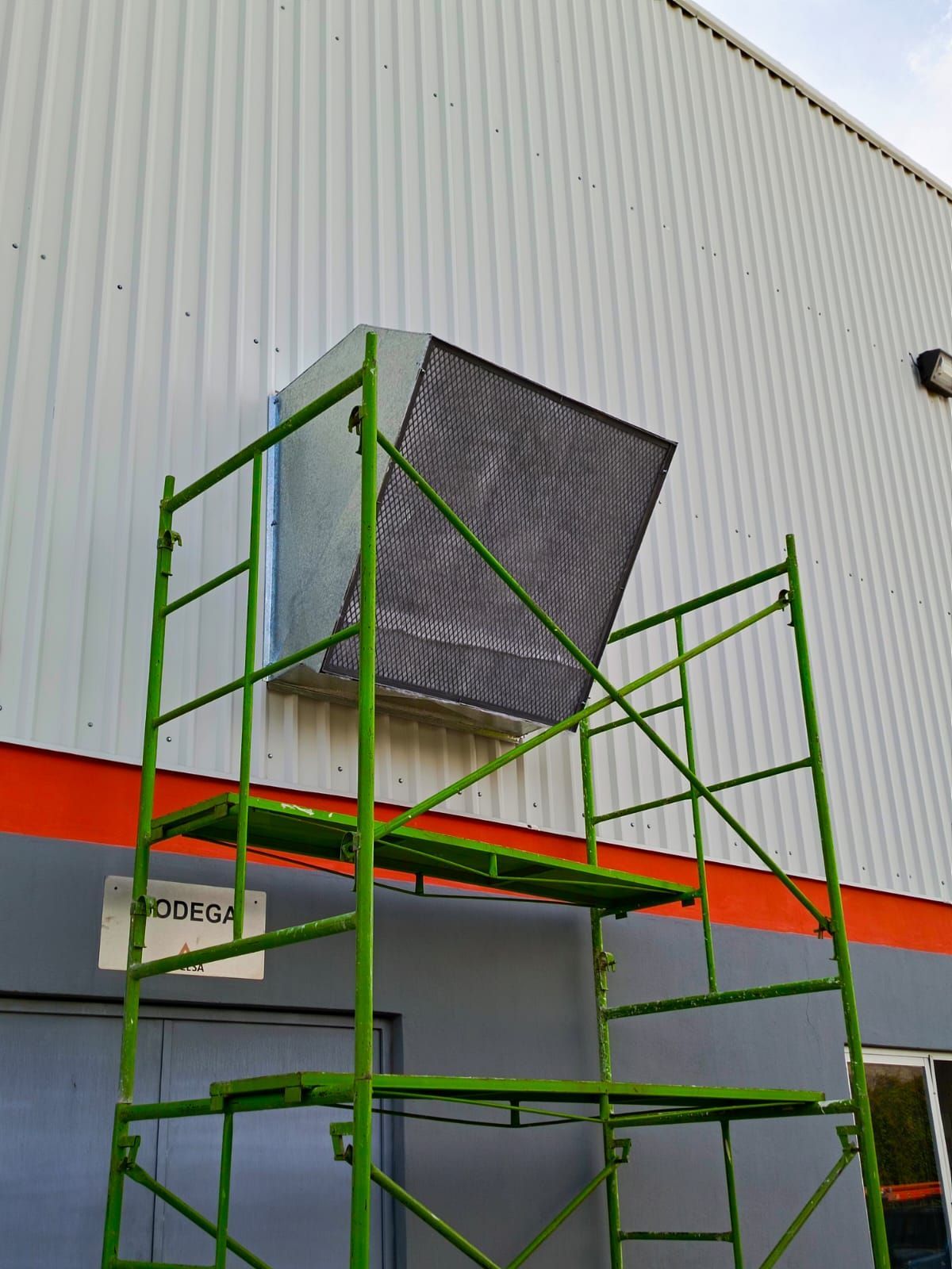 Un andamio verde se alza contra una pared, junto a una unidad de climatización instalada en el lateral de un edificio.