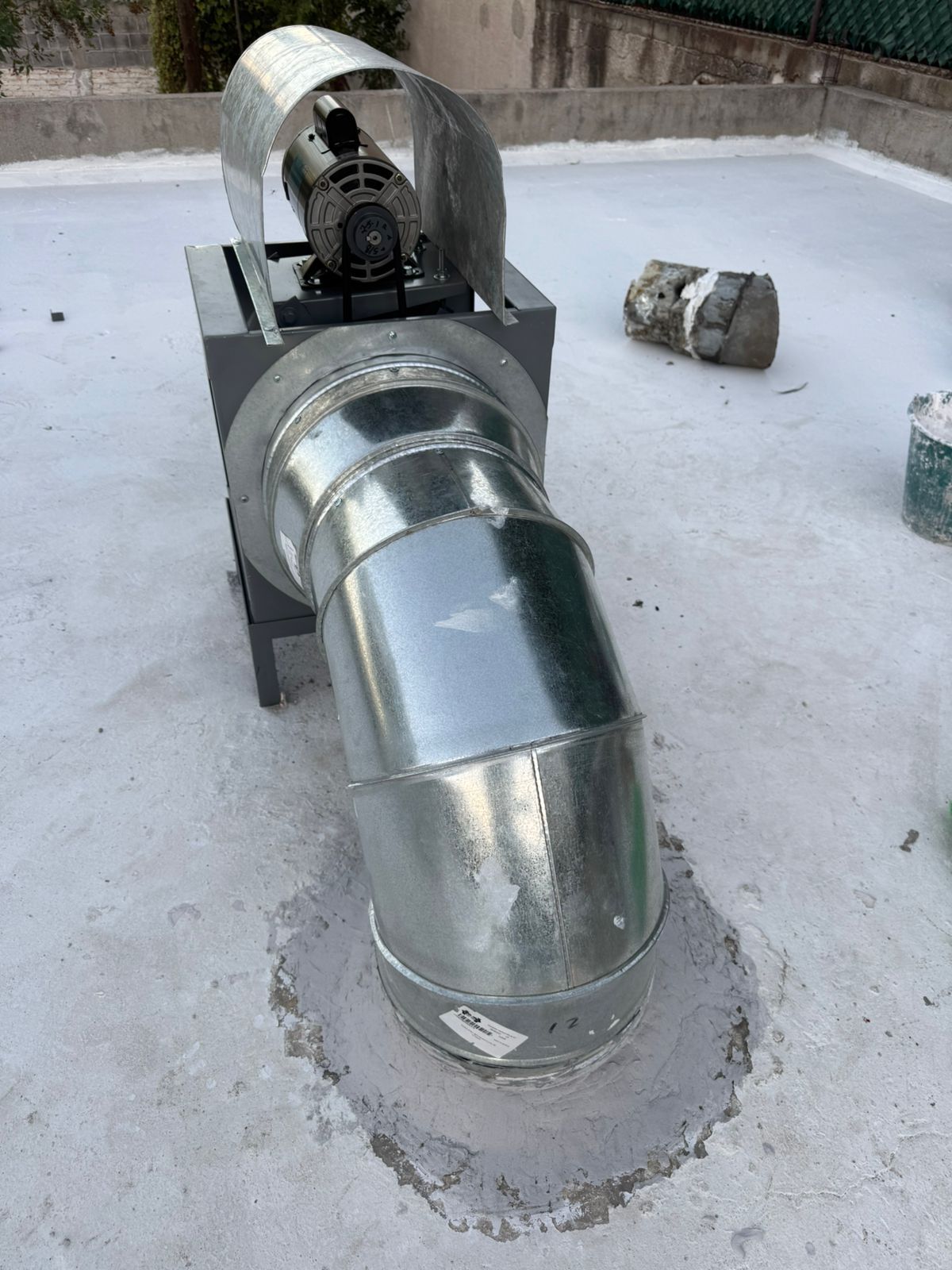 Un ventilador extractor de metal se encuentra sobre un techo plano de color gris claro, conectado a un conducto circular que atraviesa la superficie del techo.