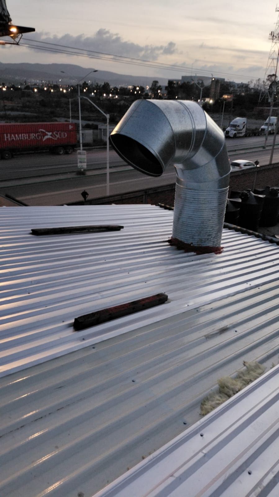 Un conducto de ventilación de metal galvanizado con la parte superior curvada, montado sobre un techo de chapa ondulada blanca al atardecer.