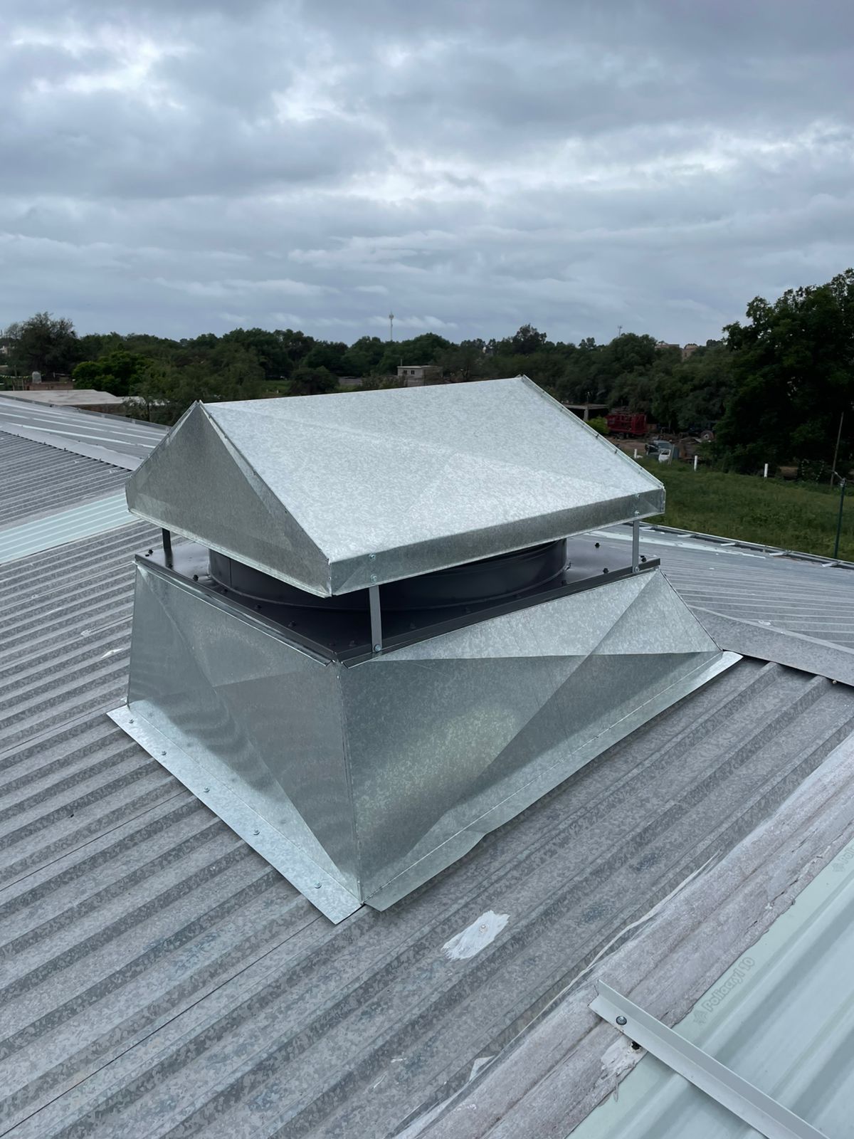Una unidad de ventilación metálica con remate a dos aguas se encuentra sobre un techo de chapa ondulada bajo un cielo nublado.