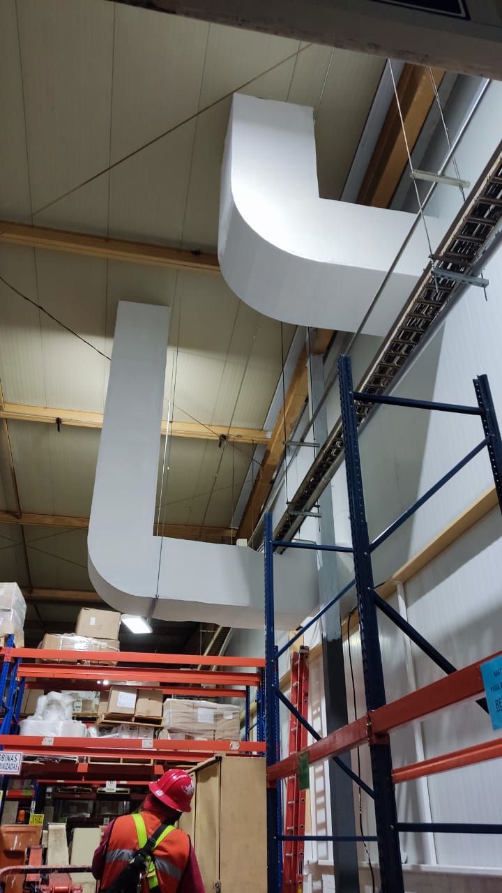 Un trabajador con ropa de alta visibilidad trabaja cerca de unos grandes conductos rectangulares blancos que cuelgan del techo de un almacén.