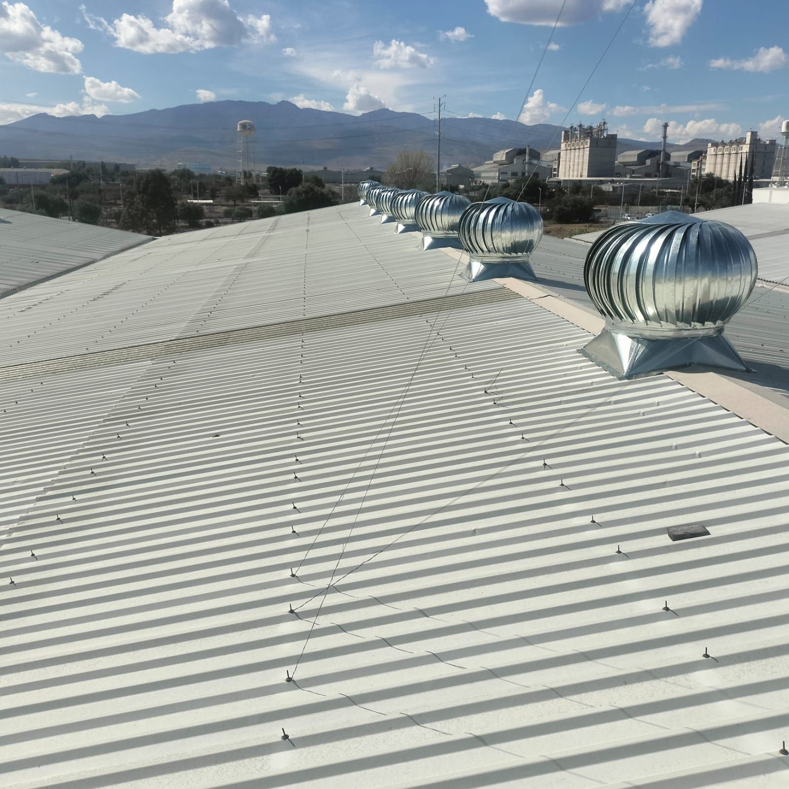 Una hilera de ventiladores de techo metálicos accionados por el viento se alza sobre un techo de chapa ondulada, con un cielo brillante y colinas a lo lejos como telón de fondo.
