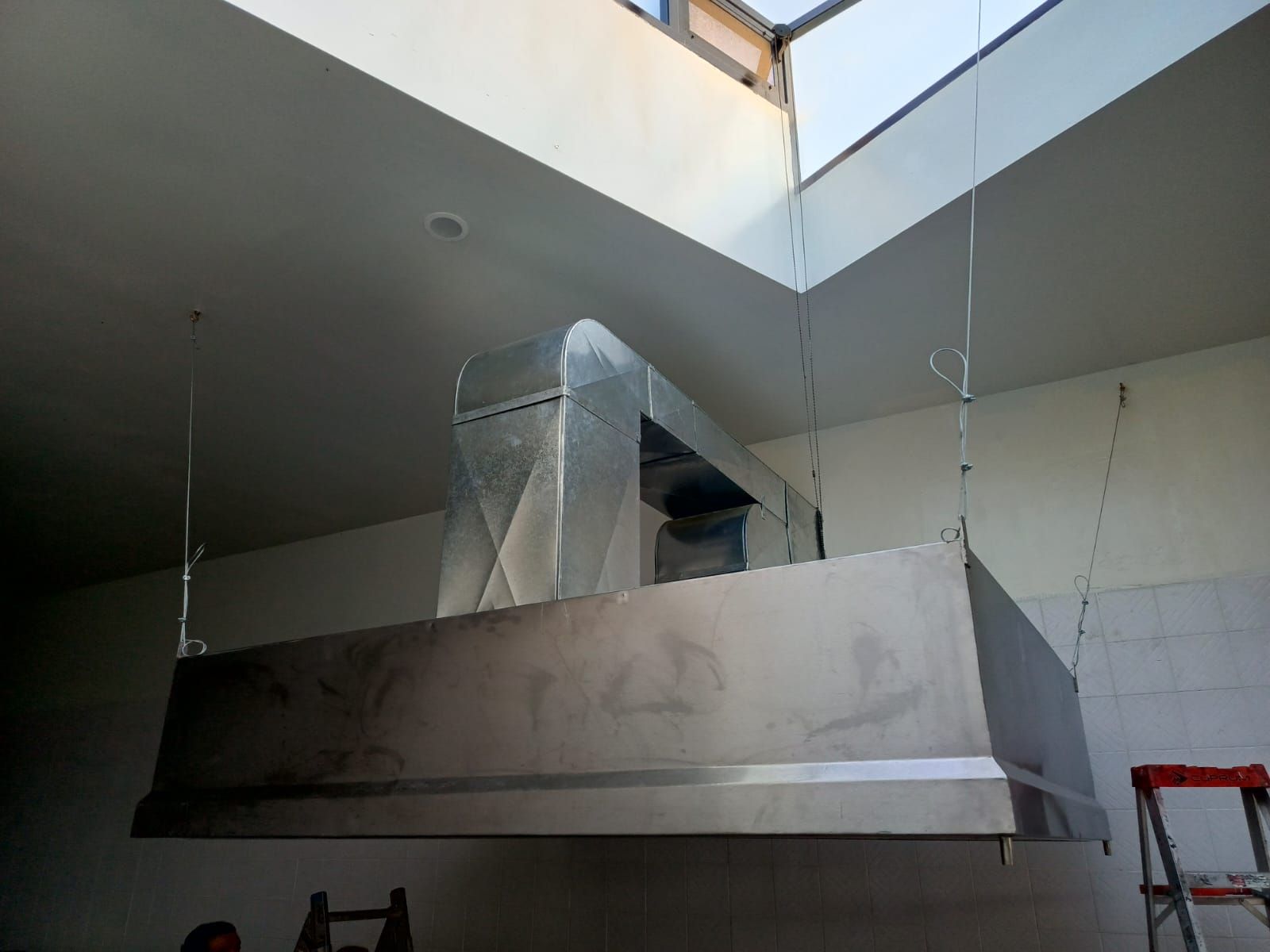 Una gran campana extractora de acero inoxidable que cuelga del techo, con un conducto metálico rectangular que se extiende desde su parte superior.