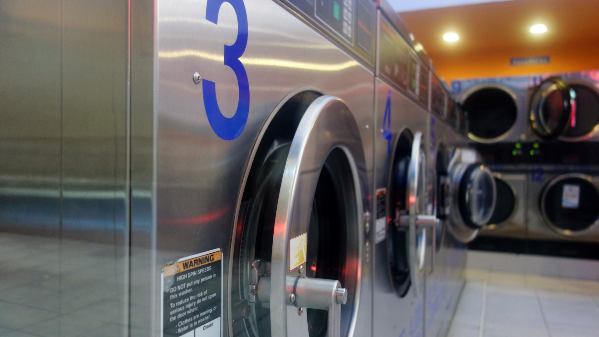 Row of stainless steel washing machines with blue numbers, in a brightly lit laundromat.