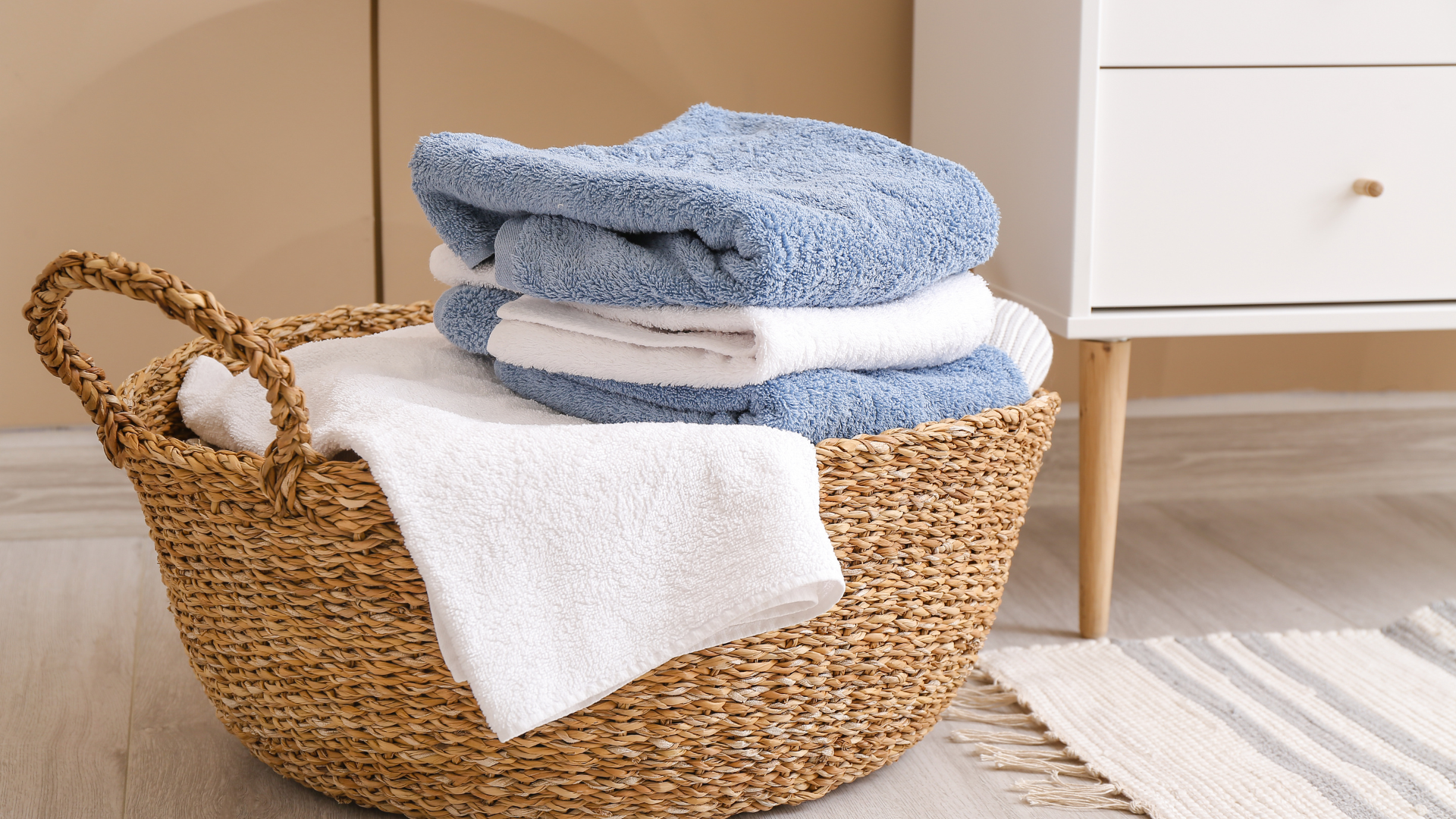 Wicker basket filled with folded white and blue towels, beside a white dresser.