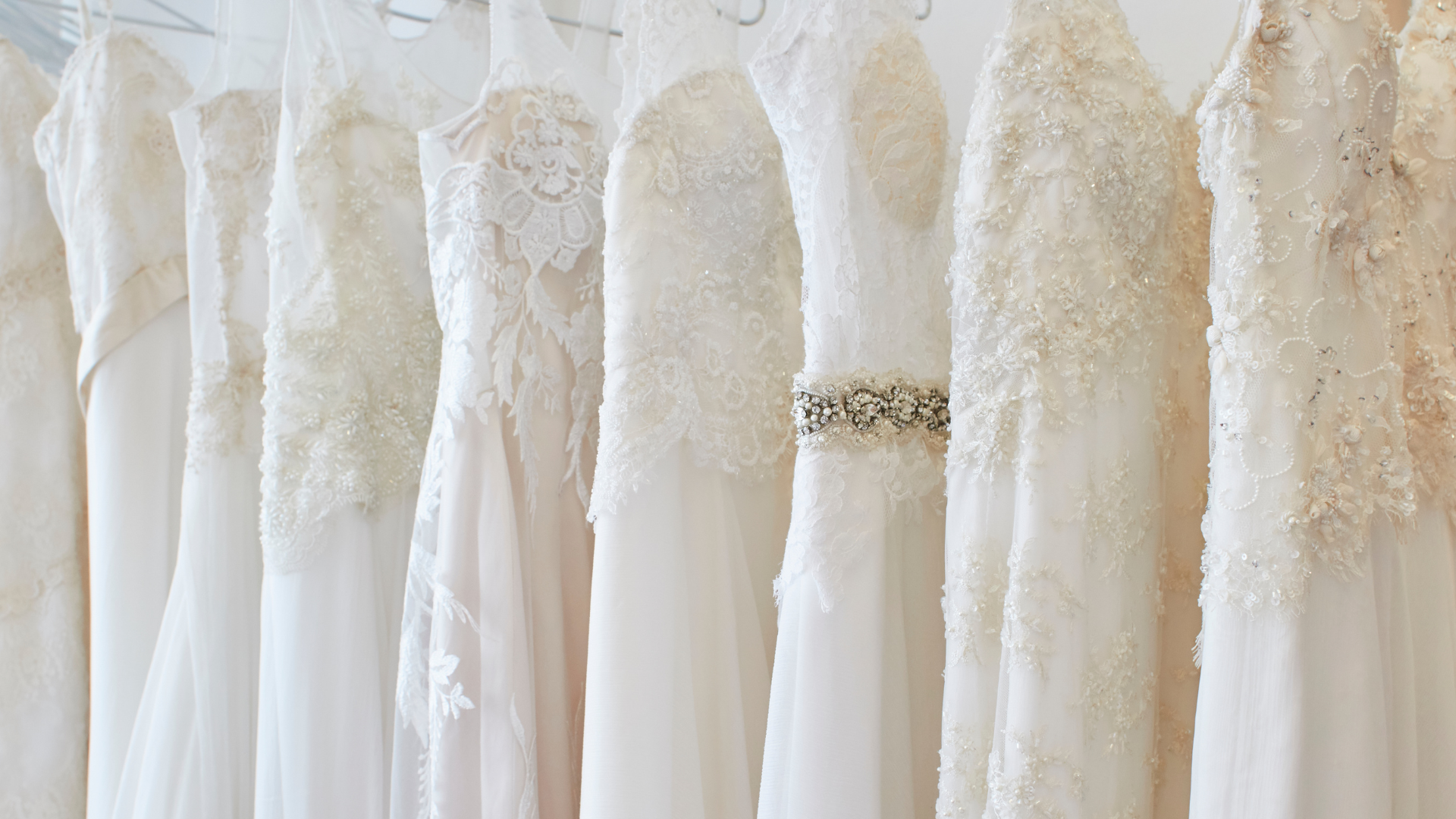 Row of white wedding dresses hanging on a rack, varying in style and embellishments.
