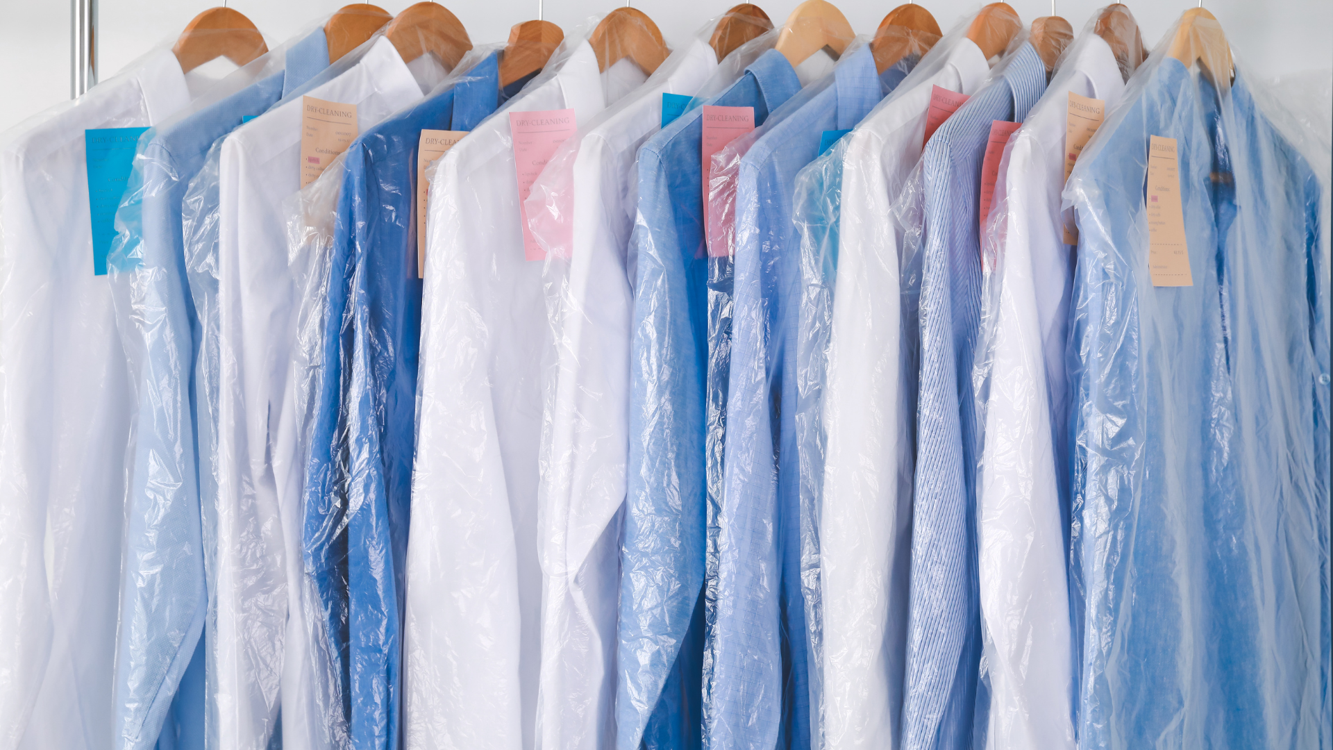 Shirts hanging in a row on wooden hangers, covered in clear plastic dry-cleaning bags. Colors include white, blue, and pink.