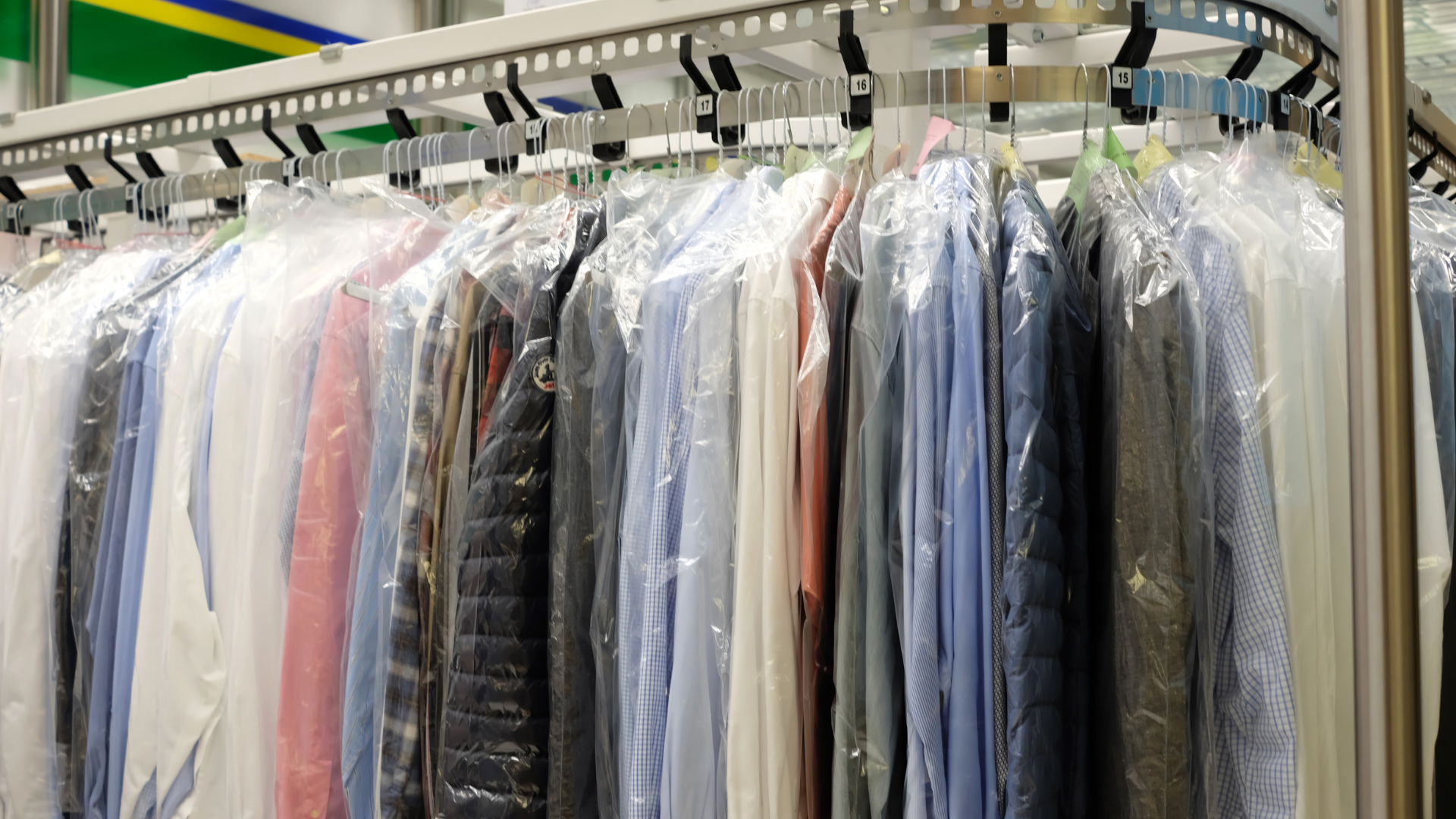 Clothes hanging in plastic dry-cleaning bags on a rack in a dry cleaner's shop. Various colors are visible.