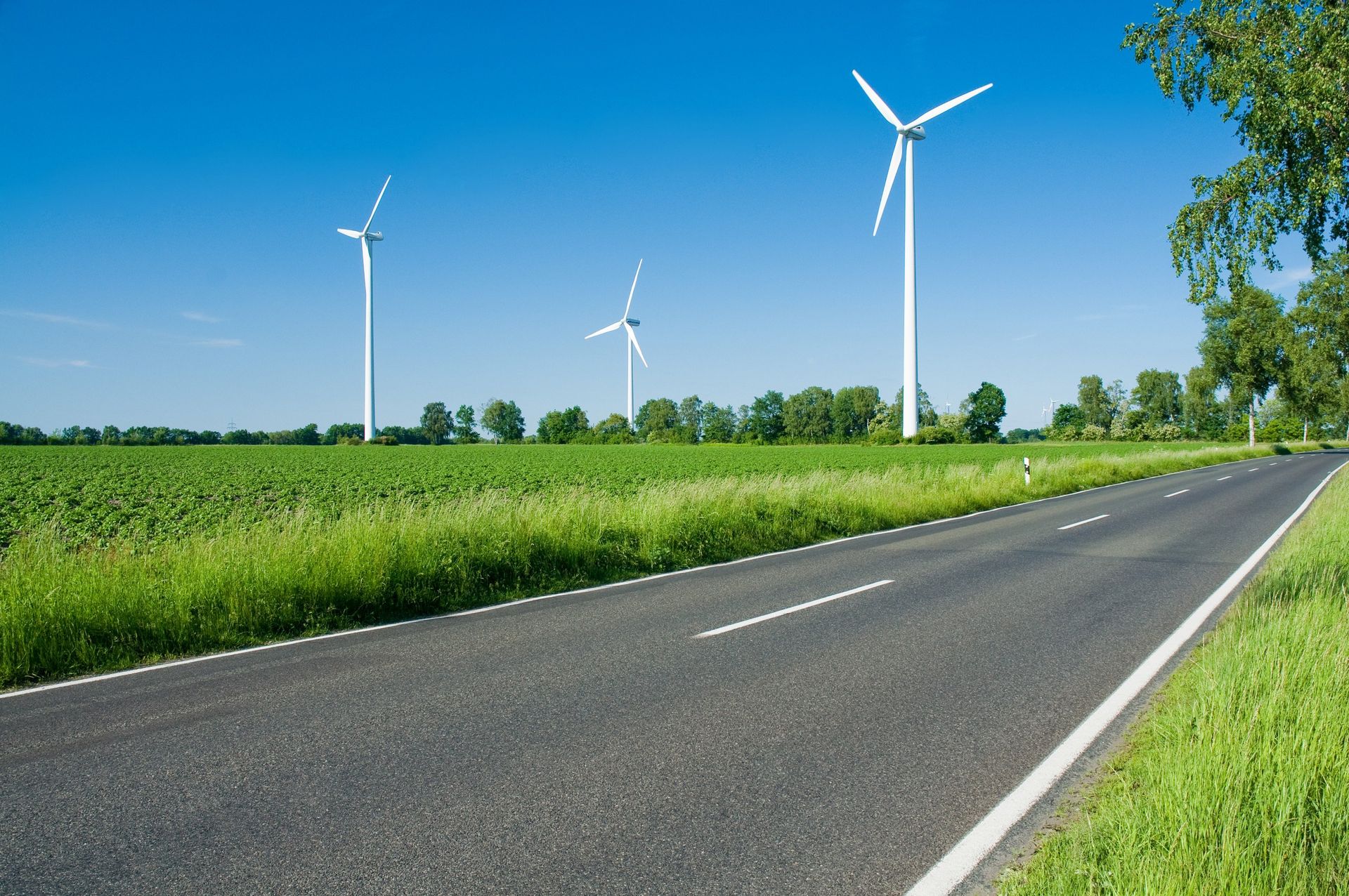 Straße entlang eines grünen Feldes mit Windkraftanlagen vor blauem Himmel.