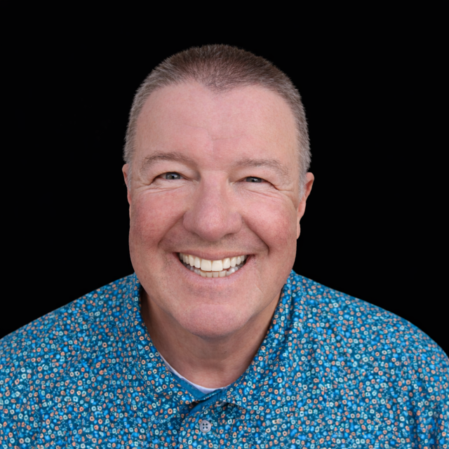 Man with a shaved head and a big smile wearing a blue patterned shirt against a black background.