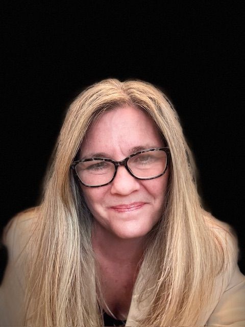 Woman with blonde hair, wearing glasses and a tan blazer, smiles against a black background.